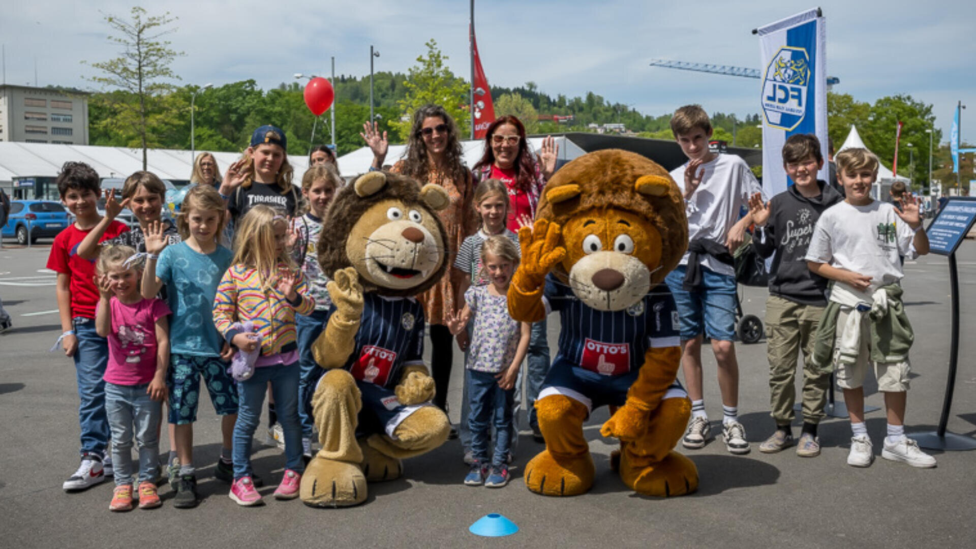Eine Gruppe von Kindern und Erwachsenen posiert im Freien, lächelnd und winkend, mit zwei Personen in Löwenkostümen. Es ist ein sonniger Tag, und im Hintergrund sind Veranstaltungsbanner, Luftballons und Gebäude zu sehen.