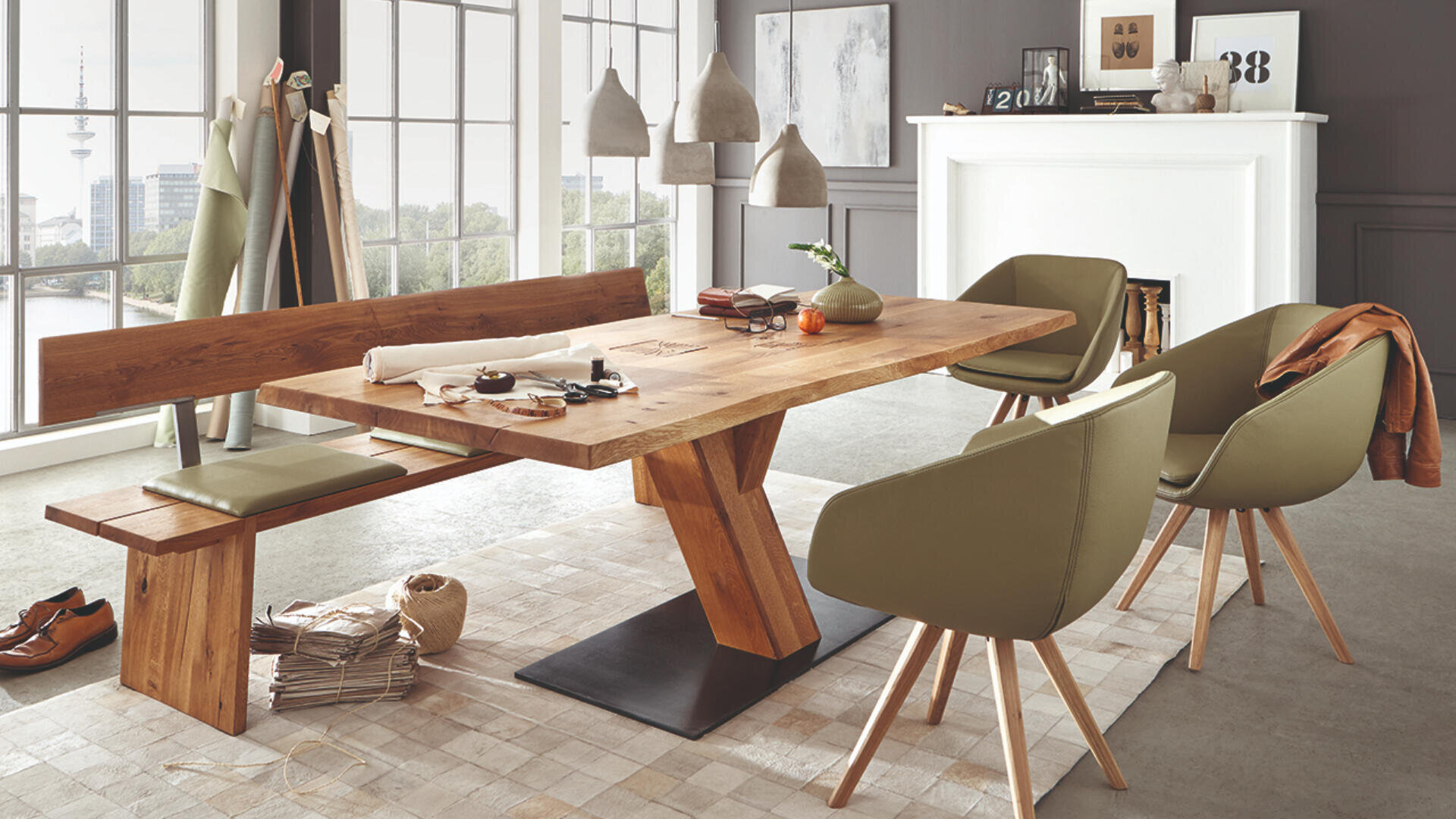 Modernes Esszimmer mit einem Holztisch, olivgrünen Stühlen und einer passenden Bank. Große Fenster lassen viel natürliches Licht herein. Dekorationsartikel und Kunst befinden sich auf einem weißen Kaminsims und Staffeleien im Hintergrund.