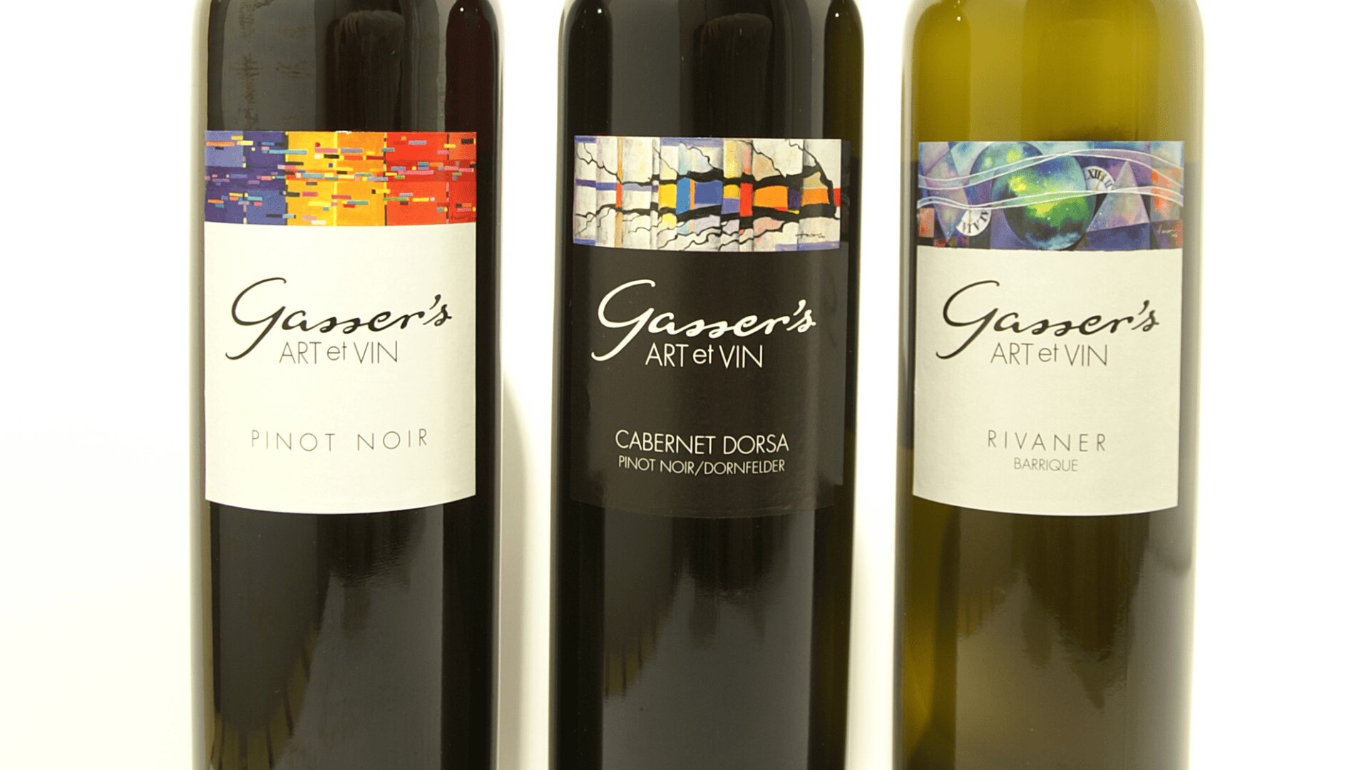 Drei Weinflaschen mit der Aufschrift "Gasser's ART et VIN" sind nebeneinander aufgestellt. Auf den Etiketten steht "Pinot Noir", "Cabernet Dorsa" und "Rivaner". Jedes Etikett zeigt ein einzigartiges abstraktes Kunstwerk im oberen Bereich.