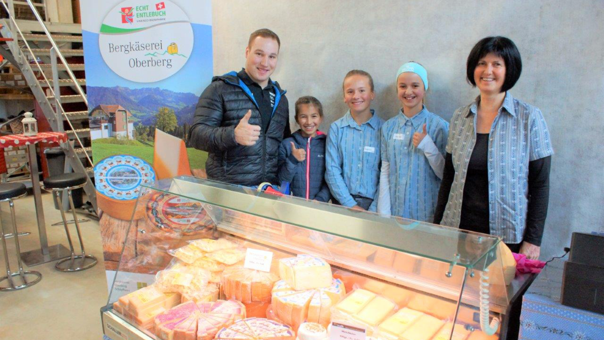 Ein Mann und vier Frauen, drei davon in passenden blauen Uniformen, stehen hinter einer Käsetheke, lächeln und zeigen die Daumen nach oben. Auf der Theke stehen verschiedene Käsesorten, und auf einem Schild steht "Bergkäserei Oberiberg".