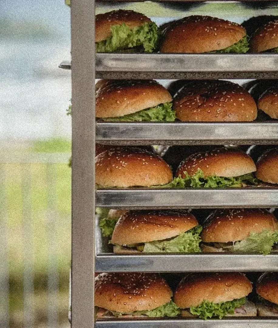 Mehrere Reihen von Sesambrötchen, gefüllt mit Salat und Patties, sind auf Metalltabletts in einem Regal gestapelt, bereit zum Servieren. Der Hintergrund ist unscharf, Elemente im Freien sind sichtbar.
