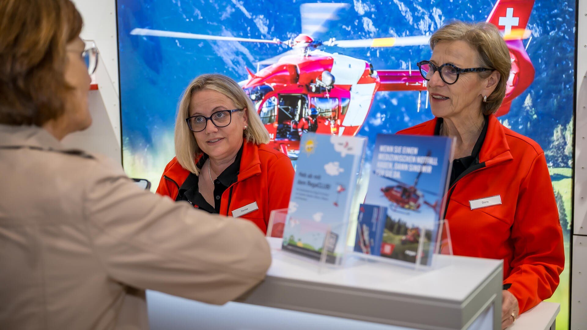 Zwei Frauen in roten Jacken stehen hinter einem Tresen und sprechen mit einem Besucher. Werbematerialien und Broschüren liegen auf dem Tresen aus. Im Hintergrund sind ein Rettungshubschrauber und Berge auf einem Bildschirm zu sehen.