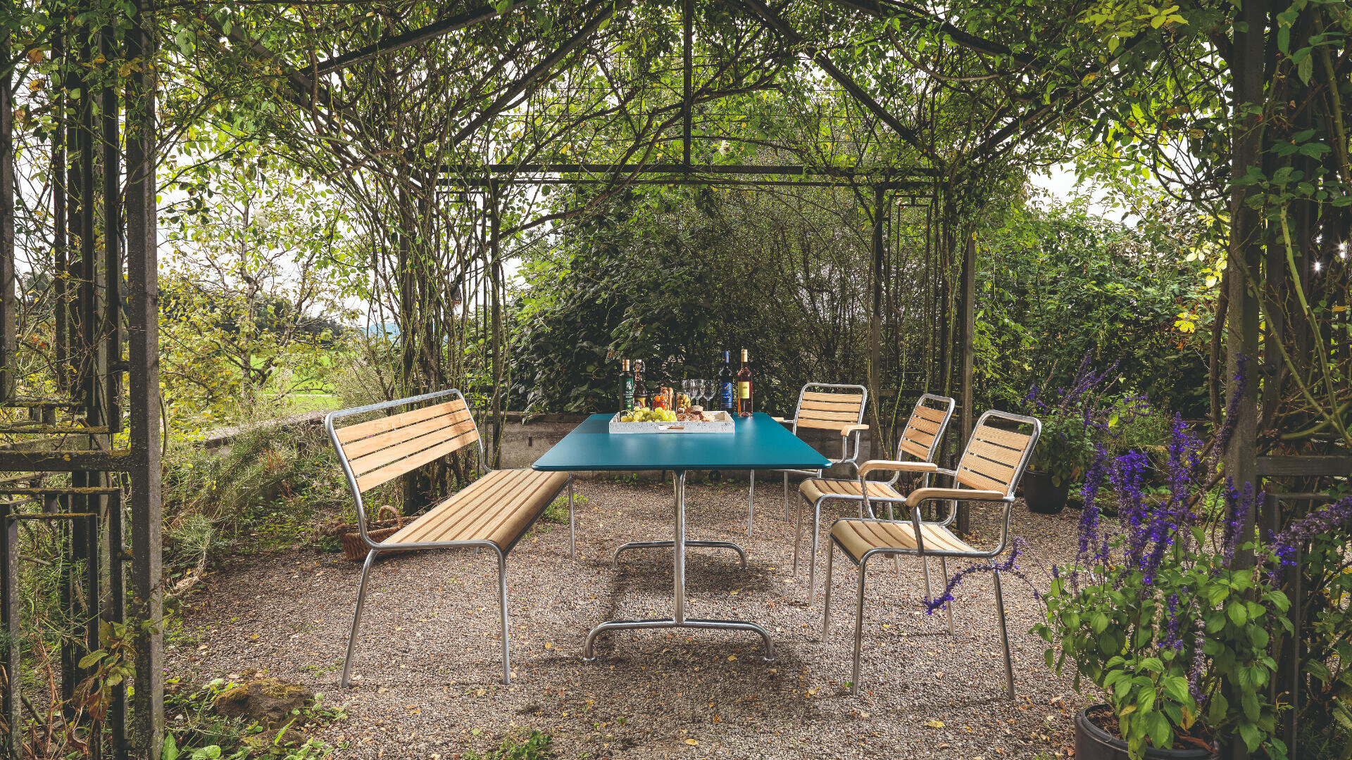 Ein Gartensitzplatz mit einem blauen Tisch, Holzbänken und Stühlen unter einer Pergola. Der Tisch ist mit Speisen, Getränken und Gläsern gedeckt, umgeben von üppigem Grün und blühenden Pflanzen auf einem Kiesboden.