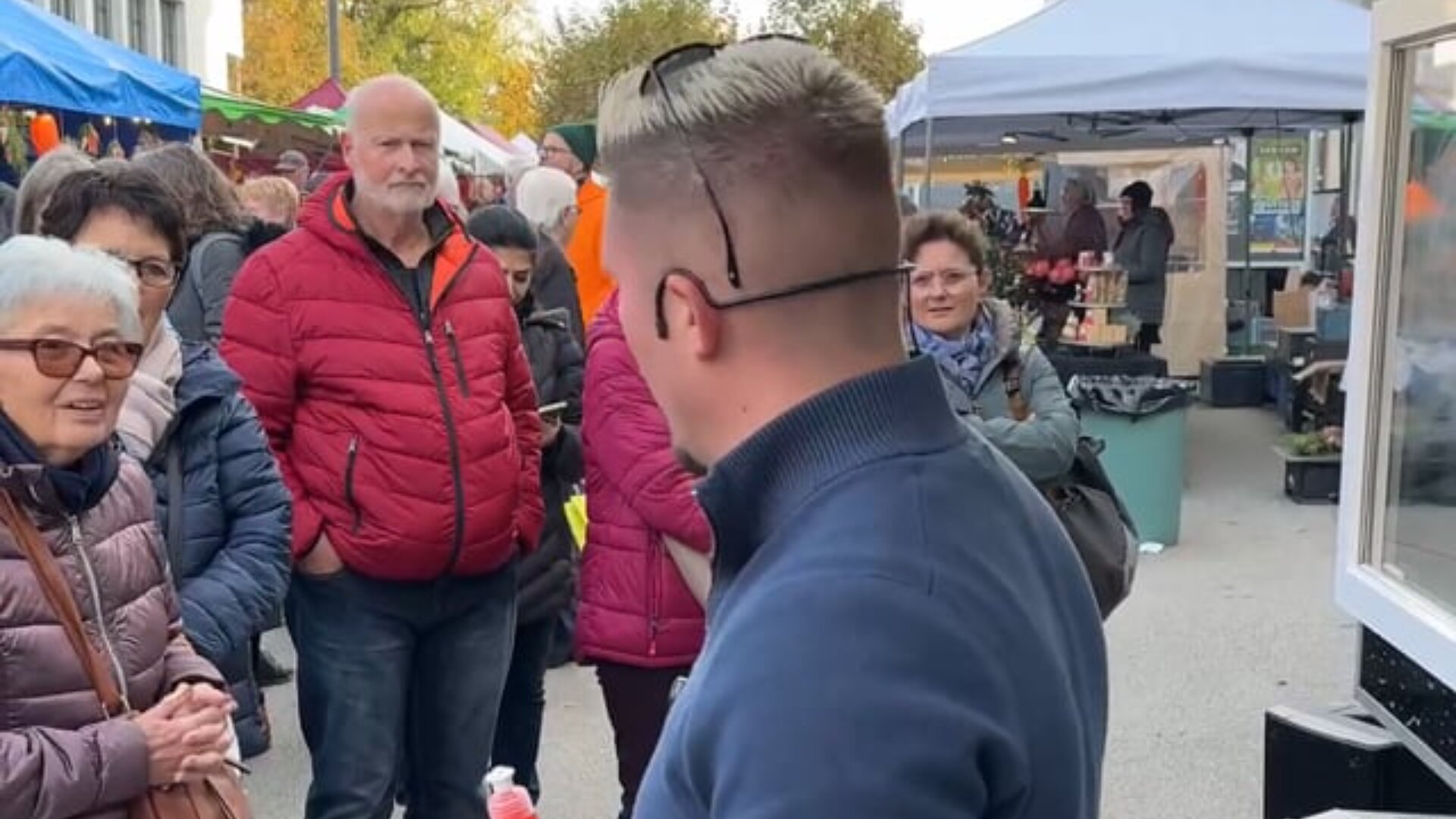 Ein Mann mit kurzen Haaren und Sonnenbrille auf dem Kopf steht einer Gruppe von Menschen auf einem Markt im Freien gegenüber, mit Zelten, Ständen und Einkäufern im Hintergrund. Einige Personen sind in Jacken gekleidet und scheinen ein Gespräch zu führen.