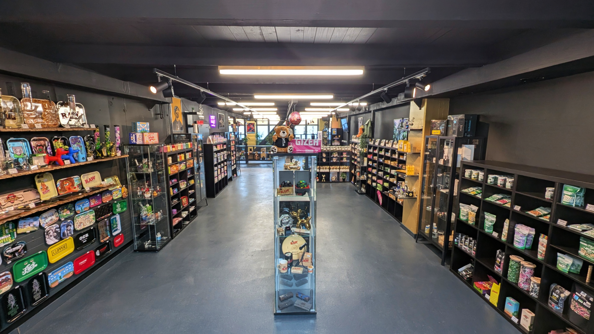 Ein gut beleuchtetes Ladeninnere mit Regalen auf beiden Seiten, in denen verschiedene Produkte und Sammlerstücke ausgestellt sind. Im Mittelgang steht eine Glasvitrine, in der die Artikel ausgestellt sind. Der Fußboden ist blau, und an der Decke befinden sich sichtbare Balken und Schienenbeleuchtung.