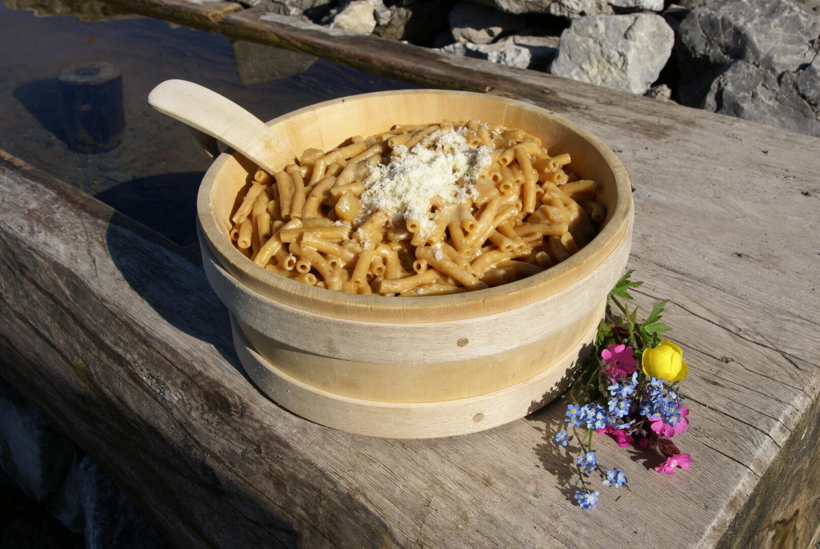 Eine Holzschüssel mit Makkaroni-Nudeln und geriebenem Käse steht auf einer rustikalen Holzfläche neben einem Strauß bunter Wildblumen und einem kleinen Teich oder Trog.