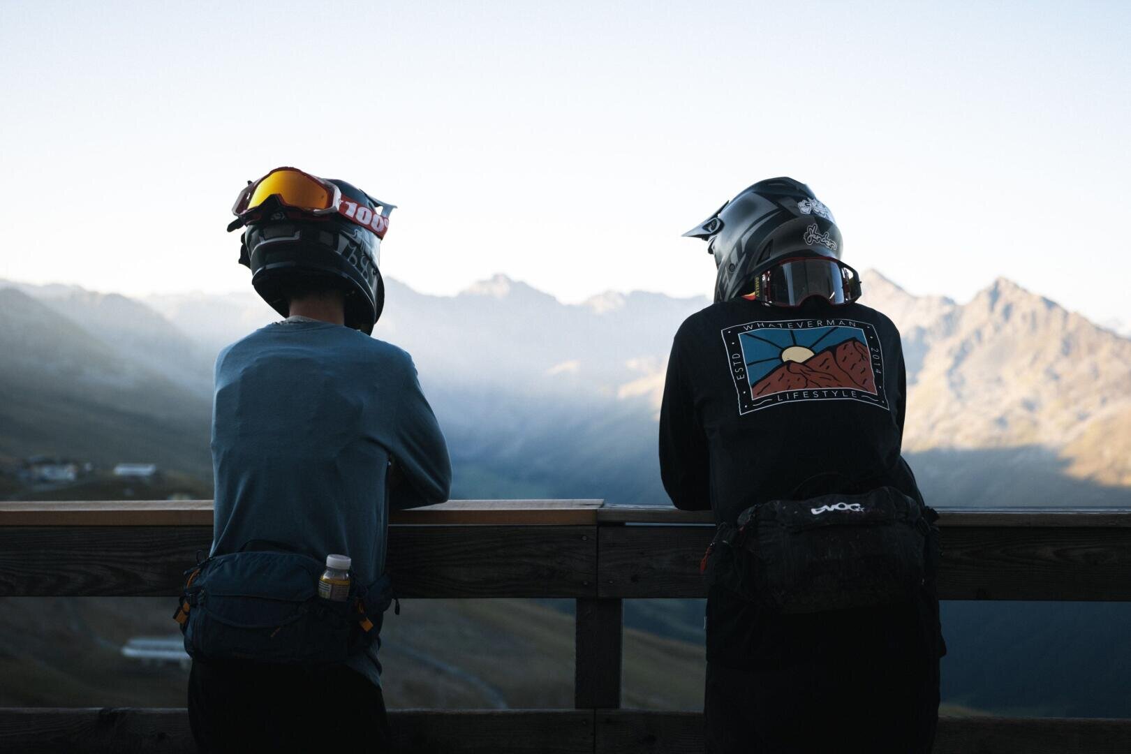 Zwei Personen mit Helmen und Sportkleidung lehnen an einem Holzgeländer und blicken auf eine malerische Berglandschaft im frühen Morgenlicht. Die Berge und Täler sind im Hintergrund sanft beleuchtet.