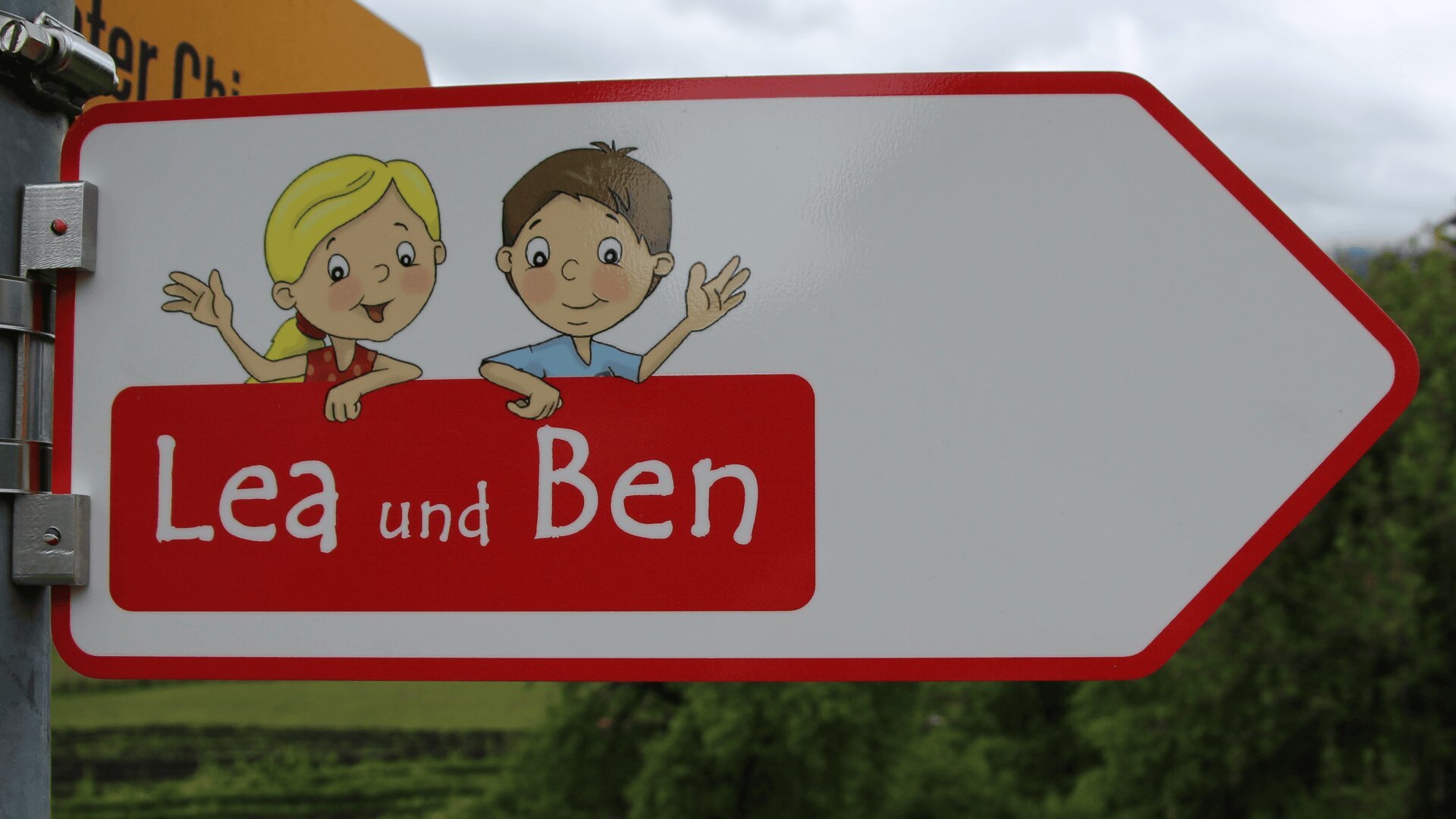 Ein weißes Pfeilschild mit winkenden Cartoon-Kindern und dem Text Lea und Ben in Rot. Der Hintergrund zeigt Grün und einen bewölkten Himmel.