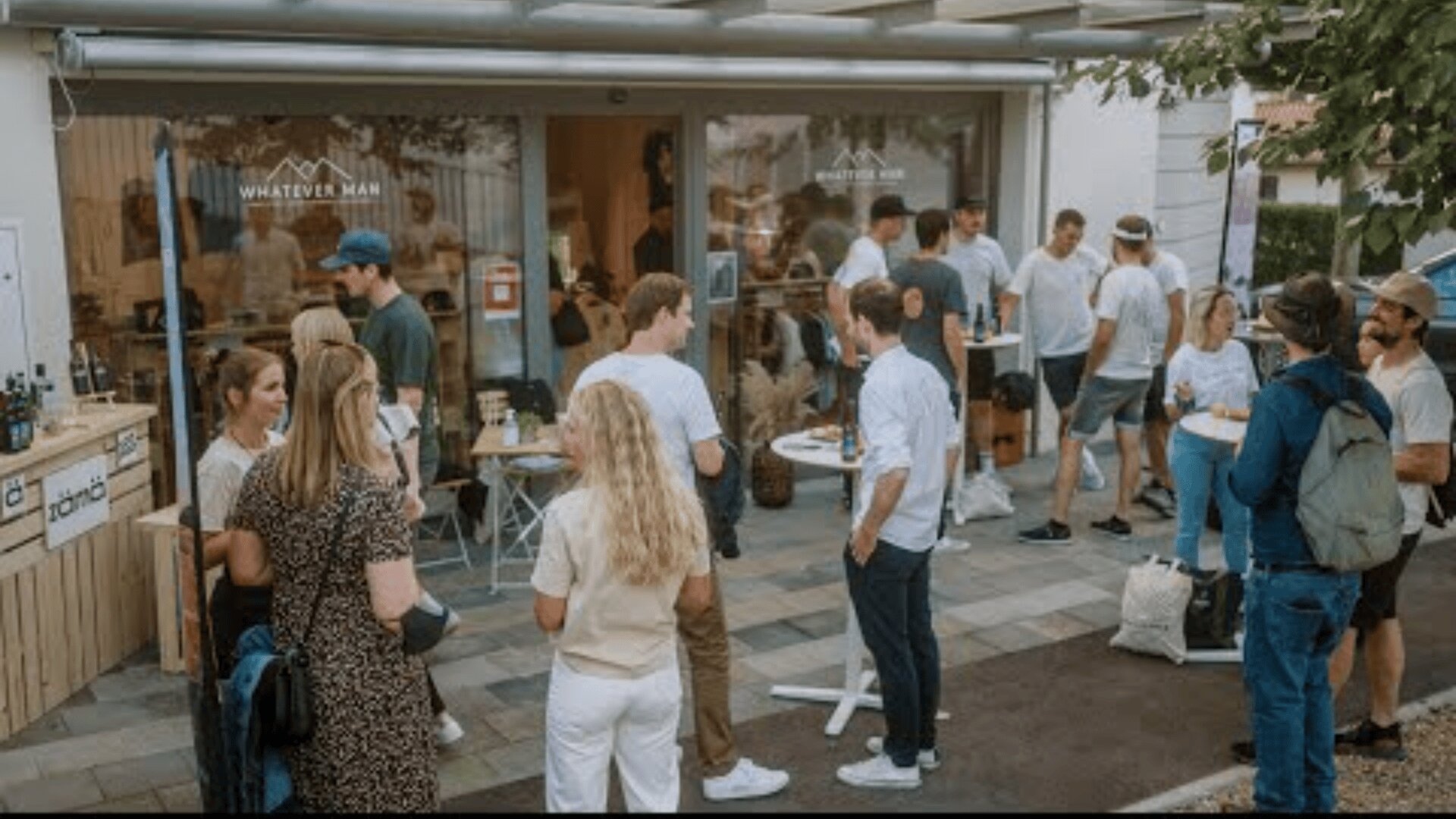 Eine Gruppe von Menschen trifft sich vor einem modernen Schaufenster mit großen Fenstern. Einige stehen in kleinen Gruppen und unterhalten sich, während andere Getränke oder Essen in der Hand halten. Die Atmosphäre ist zwanglos und entspannt, mit viel Grün und Sitzgelegenheiten im Freien.