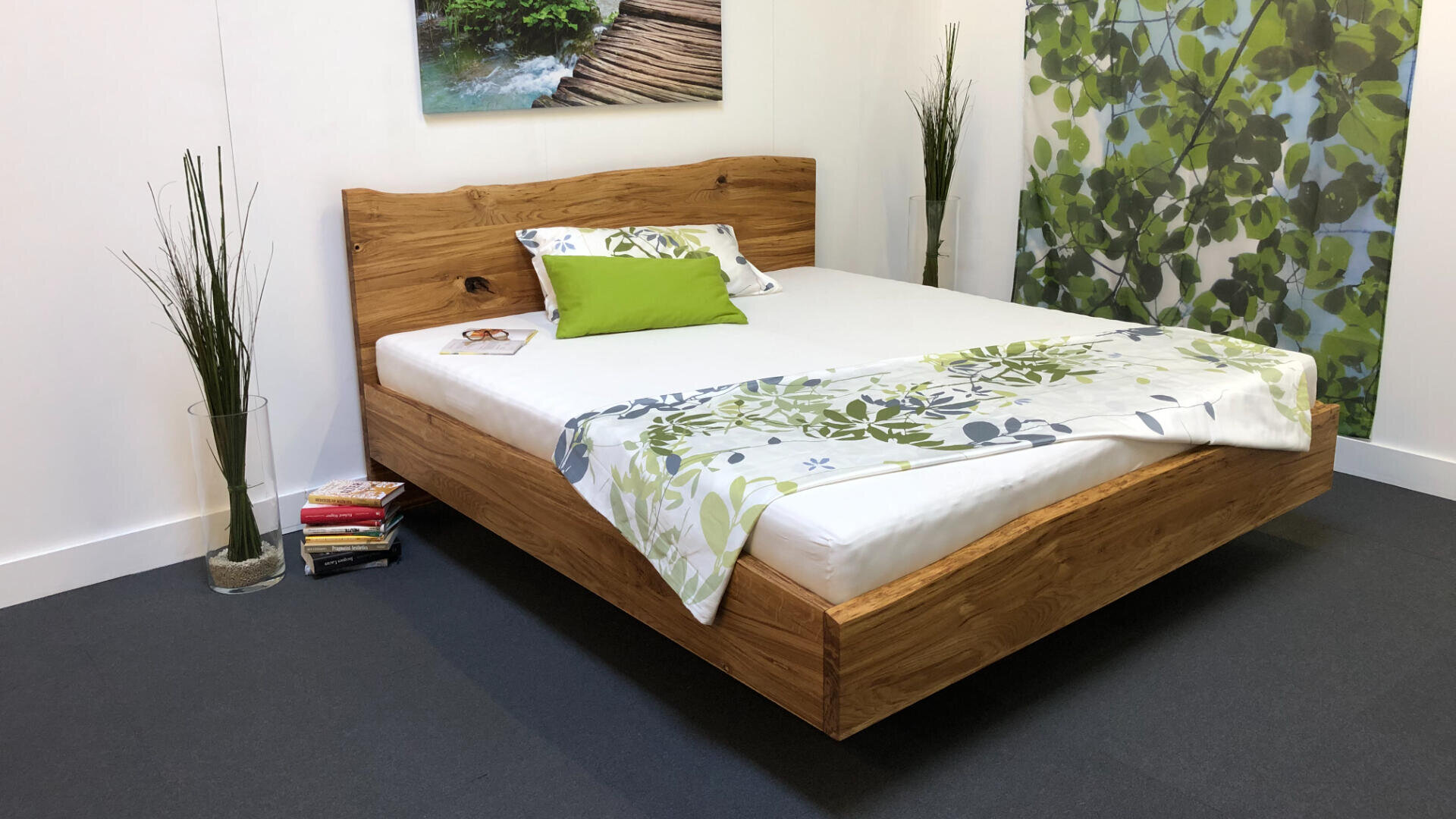 Ein modernes Schlafzimmer mit einem Bettgestell aus Holz, weißer Bettwäsche und grünen und floralen Akzenten. Ein kleiner Bücherstapel liegt auf dem Boden, neben dem Bett stehen hohe Glasvasen mit Grünzeug und im Hintergrund ist eine Wandmalerei mit Blättern zu sehen.
