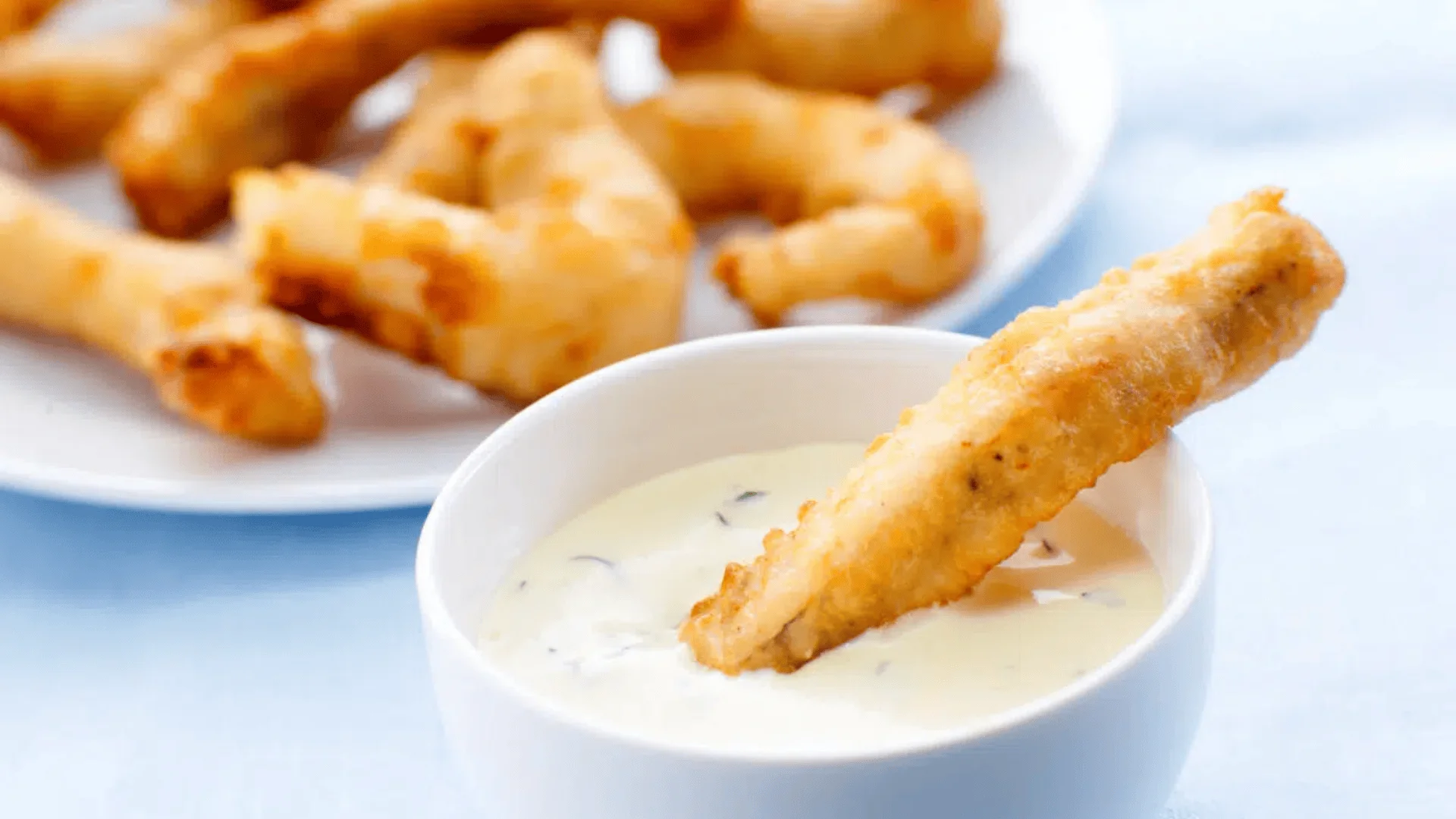 Ein knuspriger, goldener Hähnchenstreifen wird in eine Schüssel mit cremiger, weißer Dip-Sauce getaucht, während im Hintergrund weitere Hähnchenstreifen auf einem Teller liegen.