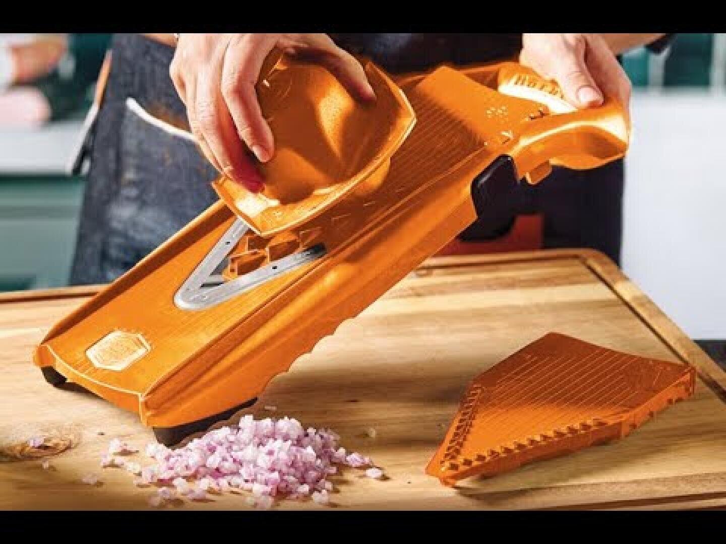 Eine Person benutzt eine orangefarbene Mandolinenschneidemaschine, um Zwiebeln auf einem hölzernen Schneidebrett fein zu hacken, wobei die gewürfelten Zwiebeln und das abnehmbare Schneideteil sichtbar sind.