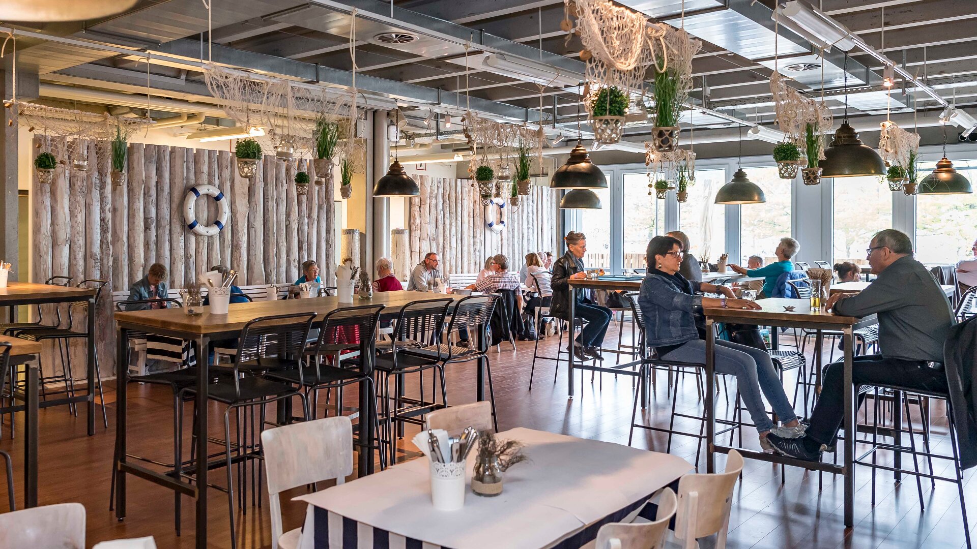 Ein geräumiges, modernes Café mit Holztischen, hohen Stühlen und Deckenpflanzen. Mehrere Personen sitzen, reden und essen in der Nähe großer Fenster, die natürliches Licht hereinlassen. Maritimes Dekor und Hängelampen schaffen eine gemütliche Atmosphäre.