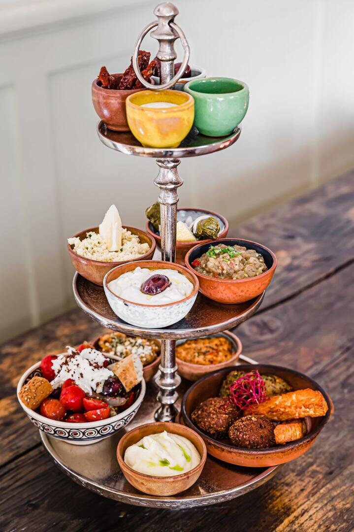 Auf einem rustikalen Holztisch stehen in einem dreistöckigen Metallständer kleine Schälchen mit verschiedenen mediterranen Vorspeisen wie Hummus, Falafel, Oliven, Traubenblätter, Dips und frisches Gemüse.