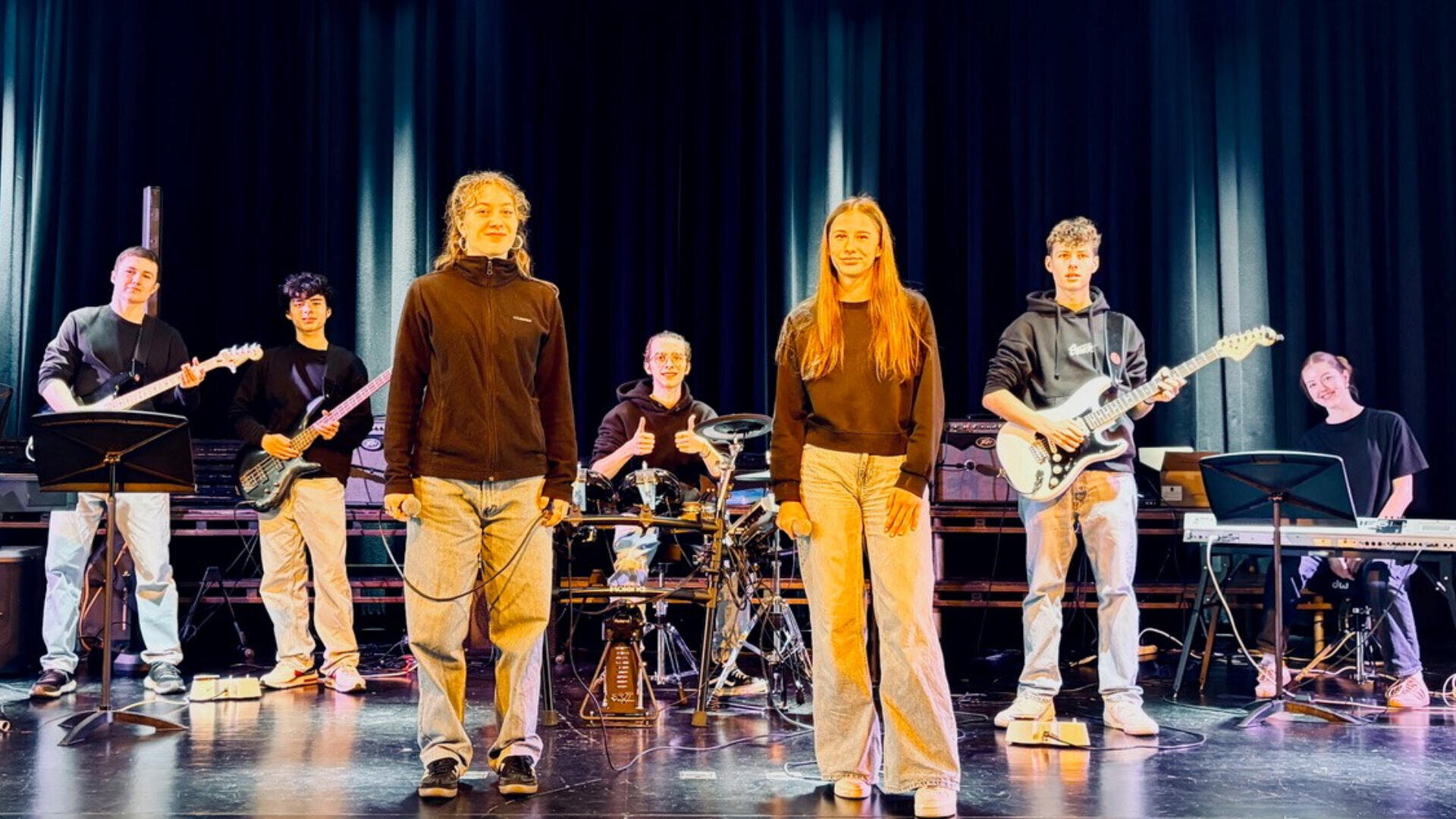 Sieben junge Musiker stehen mit Instrumenten auf der Bühne, darunter E-Gitarren, Bass, Keyboard und Schlagzeug. Zwei Sängerinnen stehen vorne und in der Mitte, alle tragen lässige schwarze Oberteile und Jeans, mit einem dunklen Vorhang im Hintergrund.
