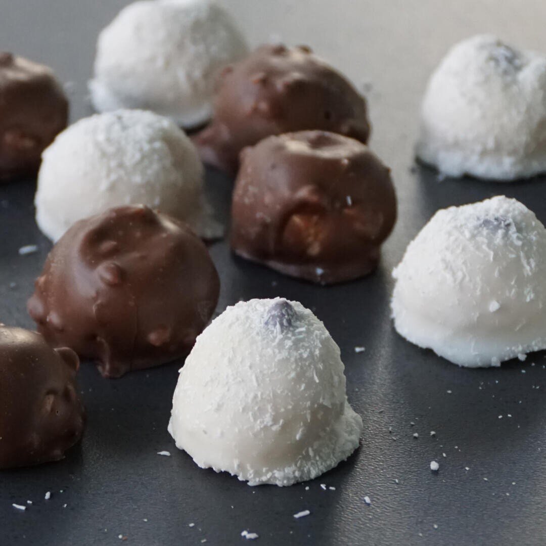 Eine Nahaufnahme verschiedener schokoladen- und weißer Kokosnussbonbons, die auf einer dunklen Fläche angeordnet sind, wobei einige Bonbons mit Schokolade und andere mit weißem Konfekt überzogen sind.