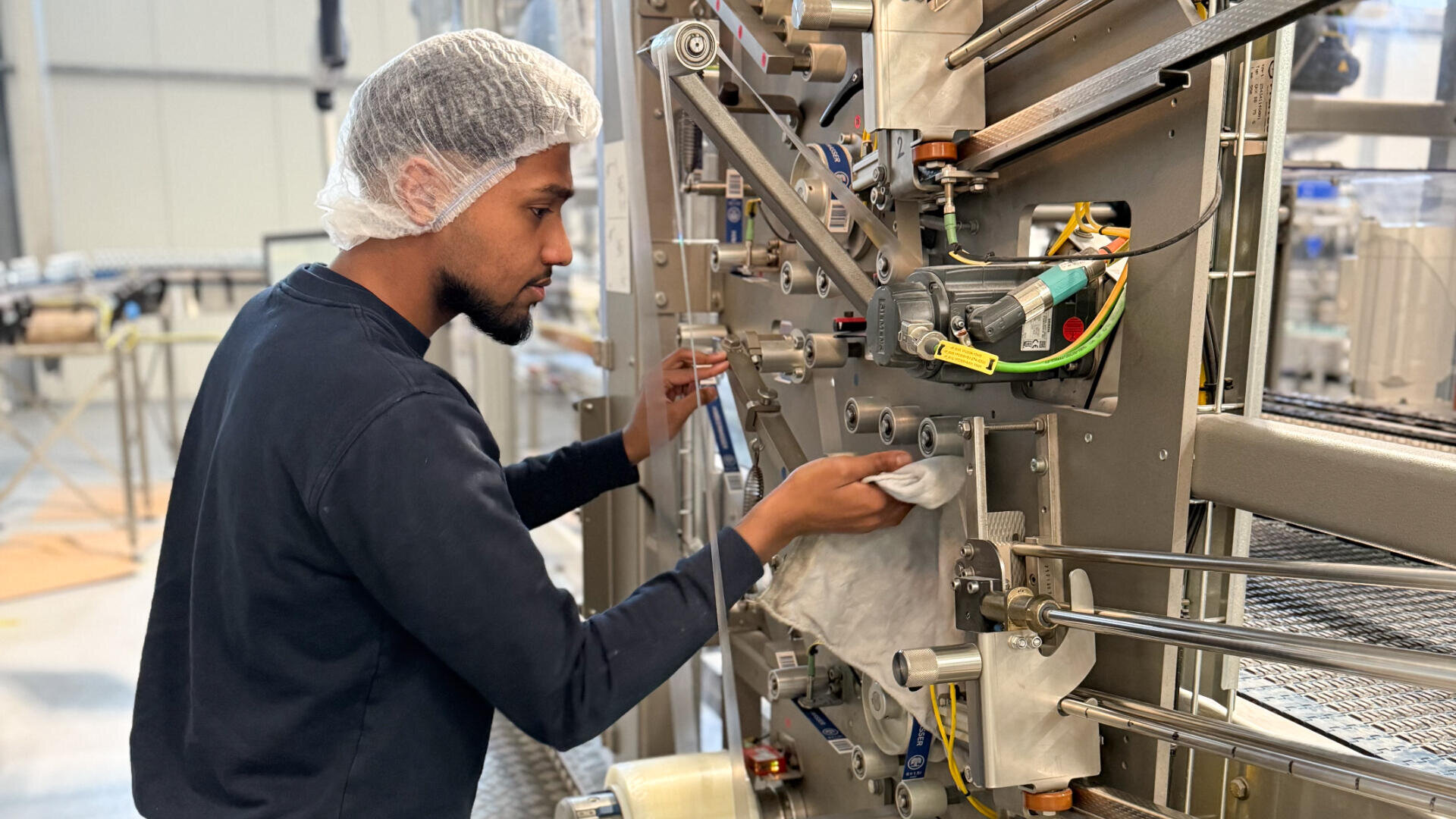Ein Arbeiter, der ein Haarnetz und ein dunkles Hemd trägt, reinigt oder wartet eine große Industriemaschine mit einem Lappen in einer Fabrikumgebung.