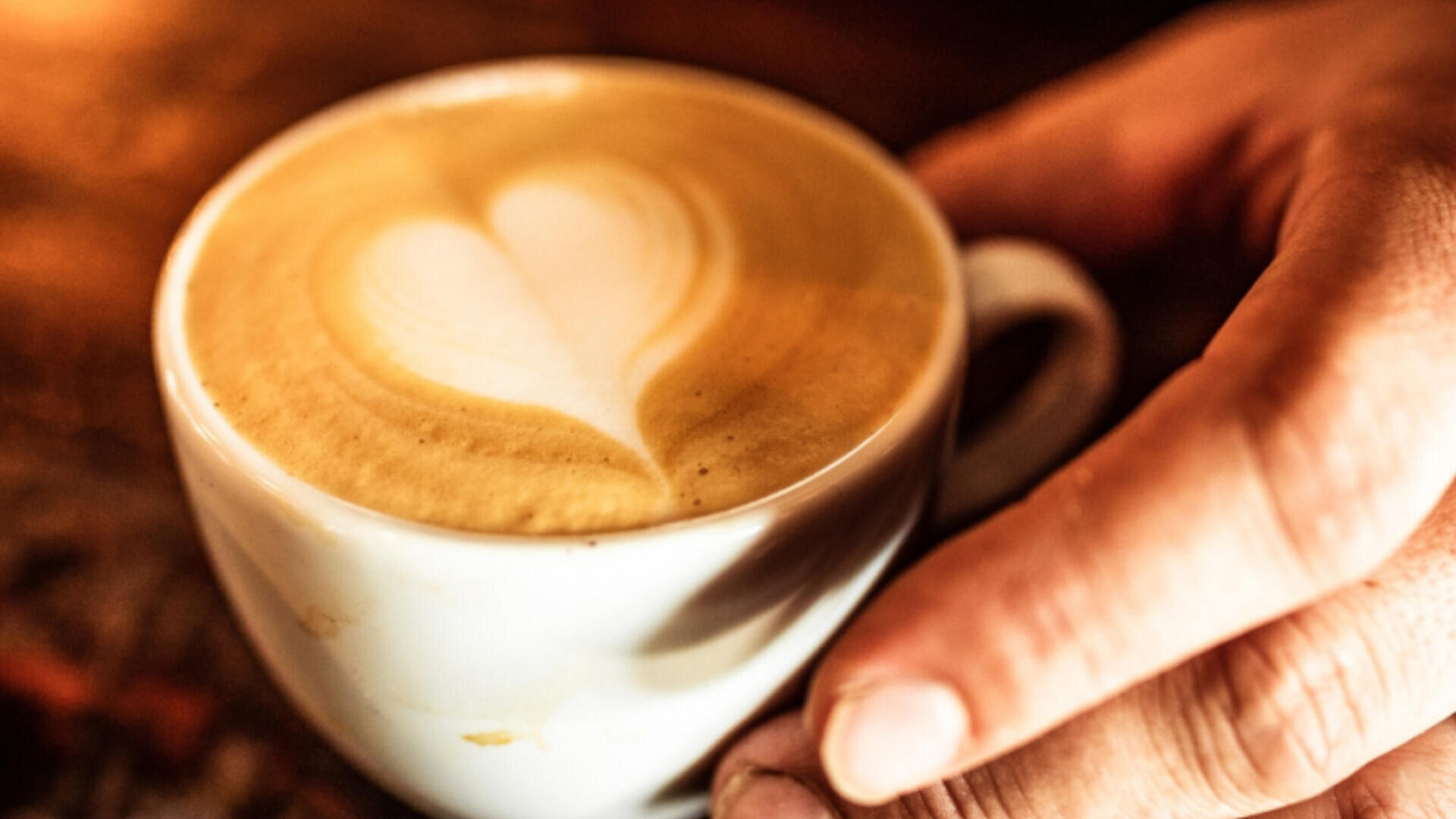 Eine Nahaufnahme einer Hand, die eine Tasse Kaffee mit einem herzförmigen Latte Art Design auf der schaumigen Oberfläche hält. Warmes, goldenes Licht schafft eine gemütliche Atmosphäre.