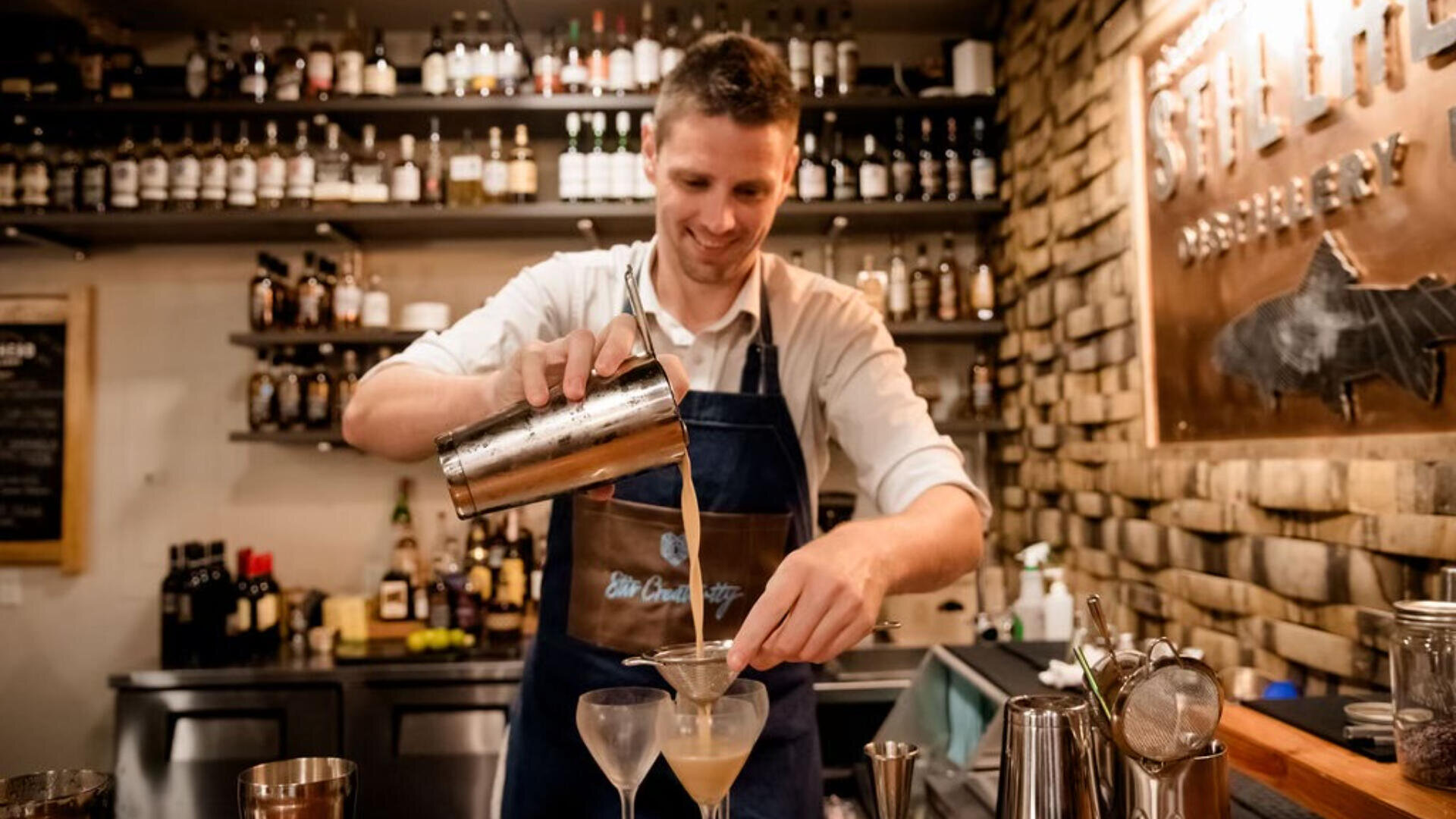 Ein Barkeeper in einer Schürze gießt lächelnd einen Cocktail durch ein Sieb in ein Glas an einer Bar, während im Hintergrund Regale mit Flaschen und Barutensilien zu sehen sind.