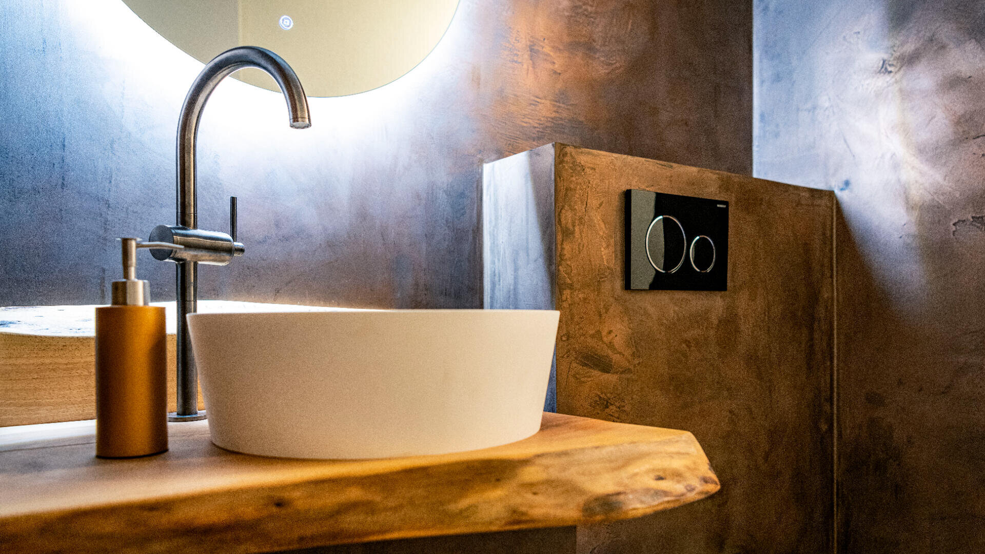 Modernes Badezimmer mit einem weißen, runden Waschbecken auf einer Arbeitsplatte aus Naturholz, einem silbernen Wasserhahn, einem goldenen Seifenspender und einem an der Wand montierten Toilettenknopf mit Zweimengenspülung vor braunen, strukturierten Wänden.