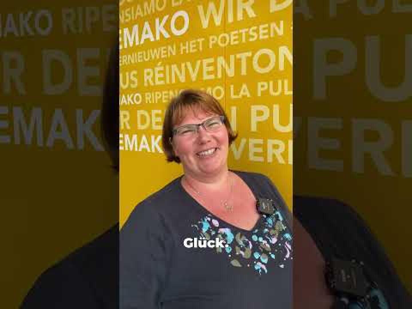 Eine lächelnde Frau mit kurzen braunen Haaren und Brille steht vor einer gelben Wand mit mehrsprachiger weißer und grauer Schrift. Neben ihrer Brust steht in weißen Buchstaben das Wort Glück geschrieben.