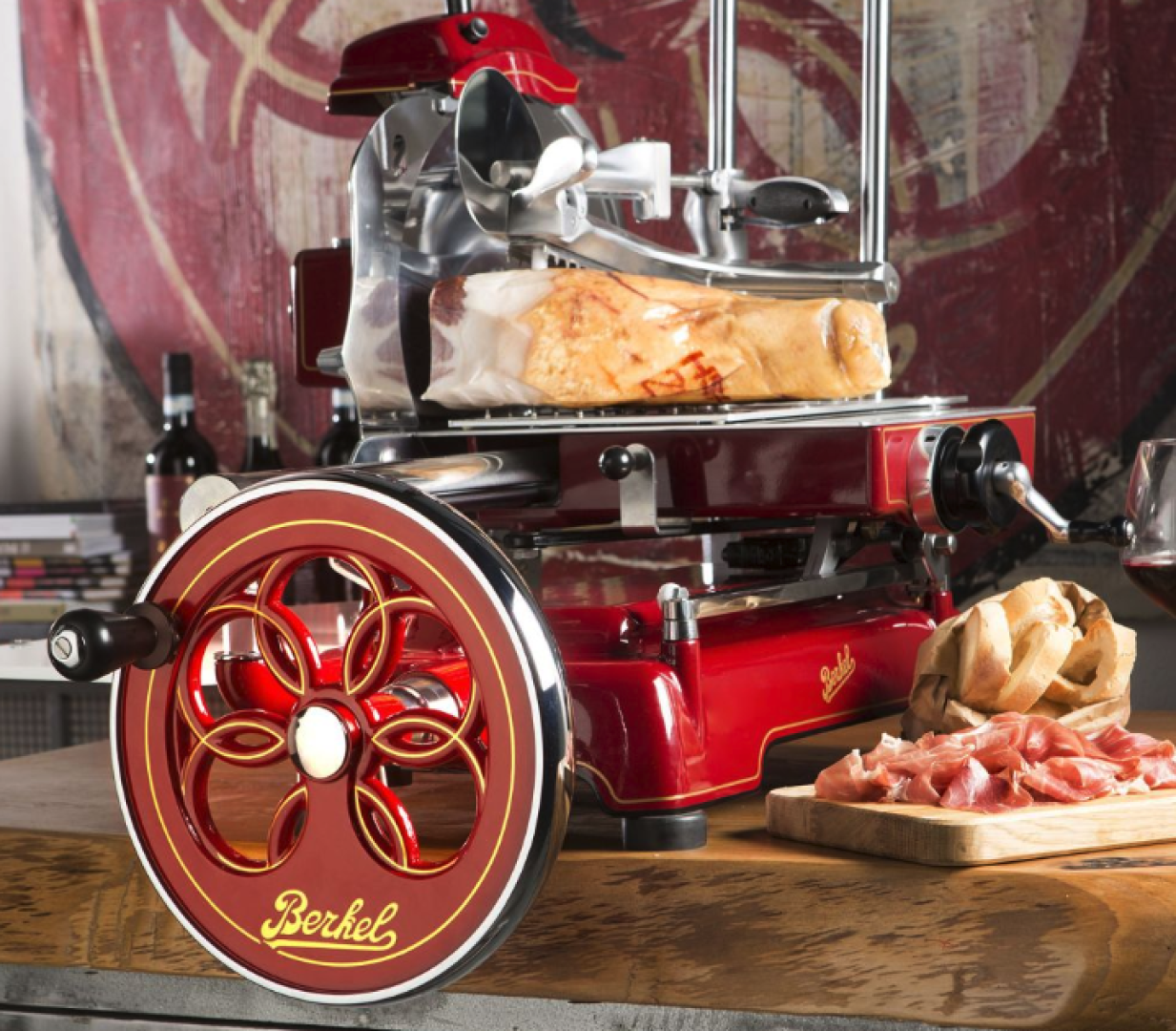 Eine alte rote Berkel-Fleischschneidemaschine hält ein großes Stück gepökeltes Fleisch. Frisch geschnittener Prosciutto und ein Baguette liegen auf einem Holzbrett daneben, im Hintergrund stehen Weinflaschen und ein Glas.