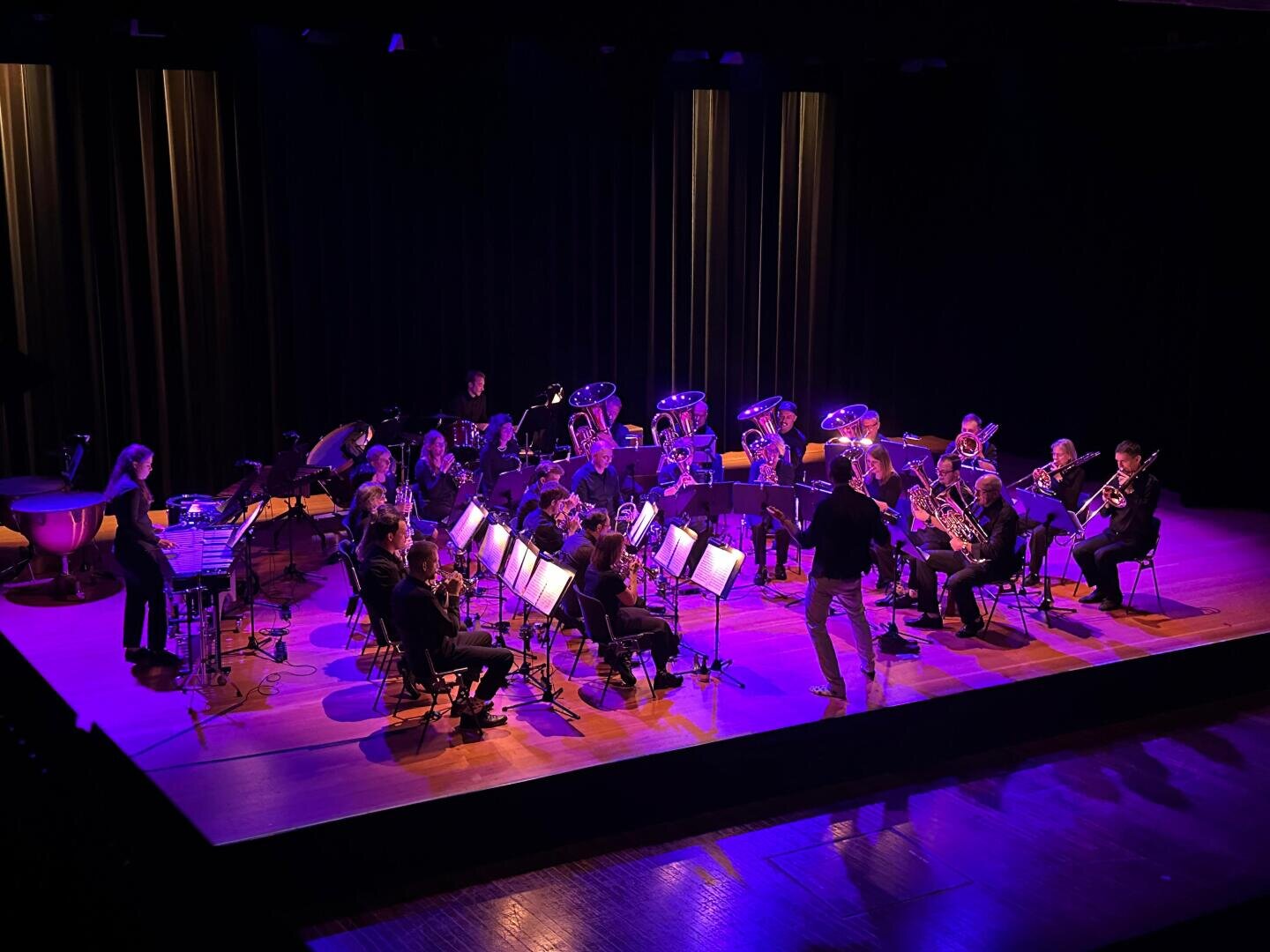 Ein Dirigent leitet ein Blech- und Holzbläserensemble auf einer mit blauem und violettem Licht beleuchteten Bühne. Die Musiker sitzen in einem Halbkreis und lesen Notenblätter, wobei verschiedene Instrumente zu sehen sind. Der Hintergrund ist dunkel mit vertikalen Vorhängen.