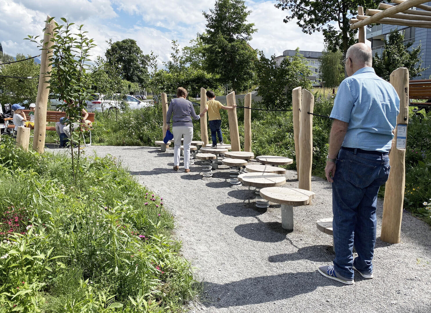 Menschen verschiedener Altersgruppen genießen einen Park mit einem Balance-Hindernisparcours mit runden, erhöhten Trittbühnen, umgeben von Grün und Wildblumen unter einem teilweise bewölkten Himmel.