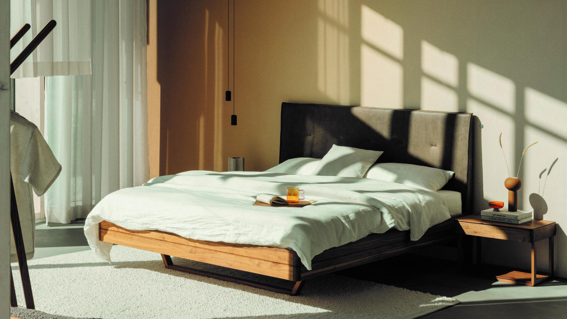 Ein sonnendurchflutetes, modernes Schlafzimmer mit einem Holzbett, weißem Bettzeug und einem dunklen, gepolsterten Kopfteil. Auf dem Bett steht ein kleiner Nachttisch mit Dekoration, einem Buch und einem Getränk, und in der Nähe der transparenten Vorhänge befindet sich ein Kleiderständer.