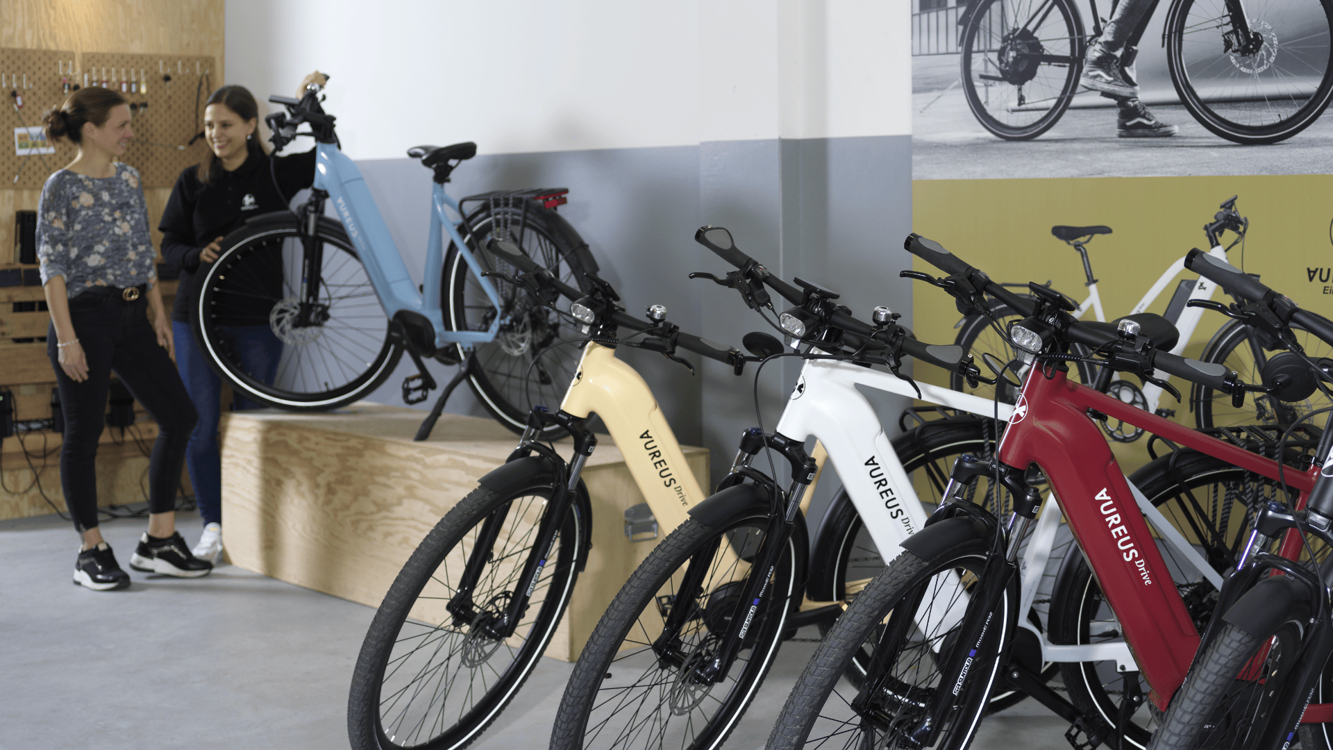Zwei Frauen stehen lächelnd neben einem hellblauen Elektrofahrrad in einem Innenraum. Im Vordergrund sind vier Elektrofahrräder in verschiedenen Farben (creme, weiß, rot, schwarz) aufgereiht. An der Wand hinter ihnen hängt ein Poster eines Fahrrads.