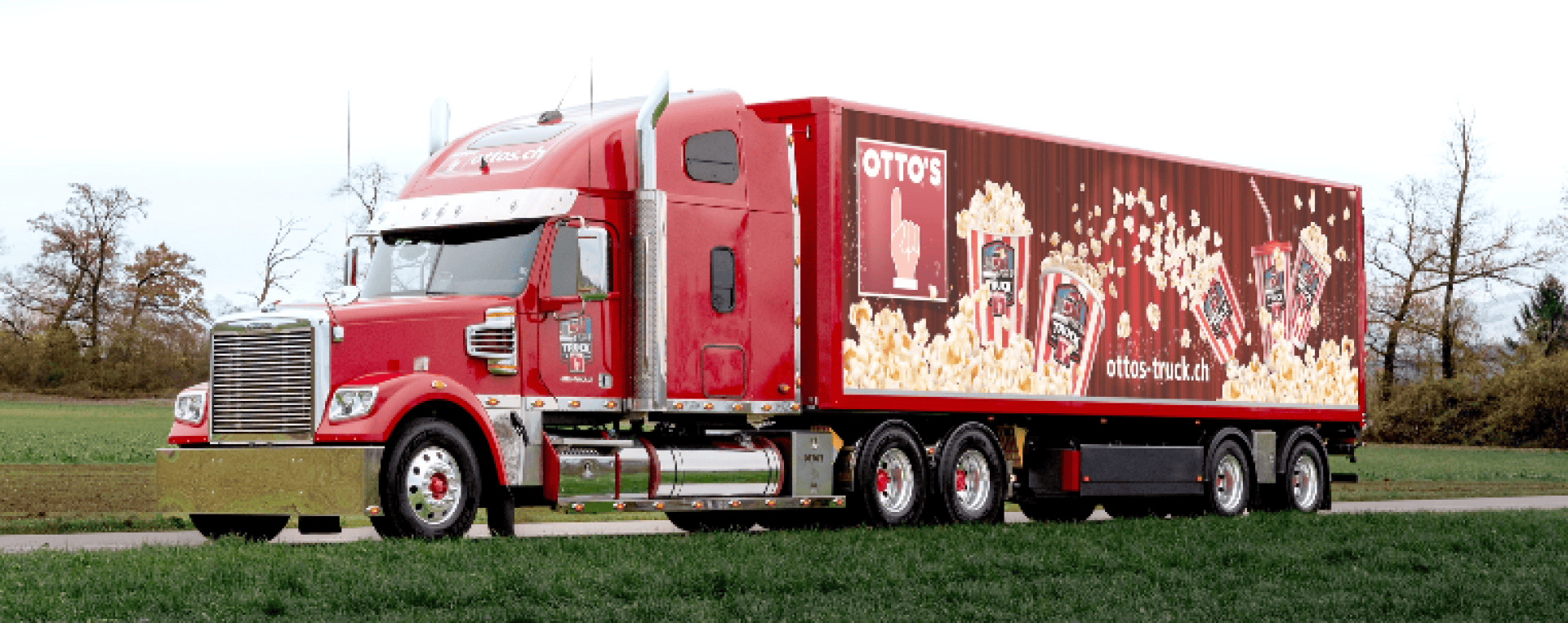 Ein roter Sattelschlepper mit einem Anhänger ist neben einem Feld geparkt. Auf dem Anhänger sind Bilder von Popcorn- und Snackpaketen mit dem Text OTTOS und hellen, farbenfrohen Grafiken an der Seite zu sehen.