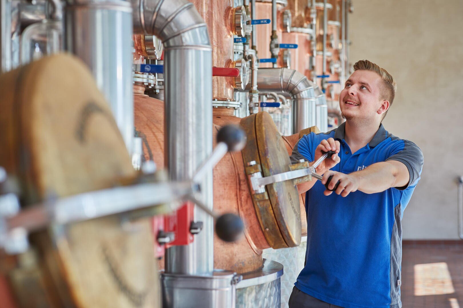Ein Mann im blauen Hemd betätigt ein Ventil an einem großen kupfernen Destillationstank in einer Brauerei oder Brennerei und lächelt, während er zwischen Metallrohren und Geräten arbeitet.