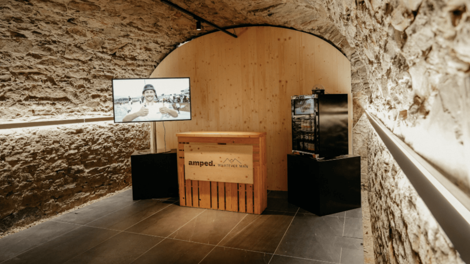 In einem kleinen, modernen Raum mit Steinwänden und einer hölzernen Rückwand stehen ein Fernseher, auf dem ein lächelnder Mann zu sehen ist, ein Schreibtisch aus Holzkisten mit der Aufschrift amped. und ein schwarzer Minikühlschrank auf einem Ständer. Eine warme Beleuchtung verläuft an der Wand entlang.