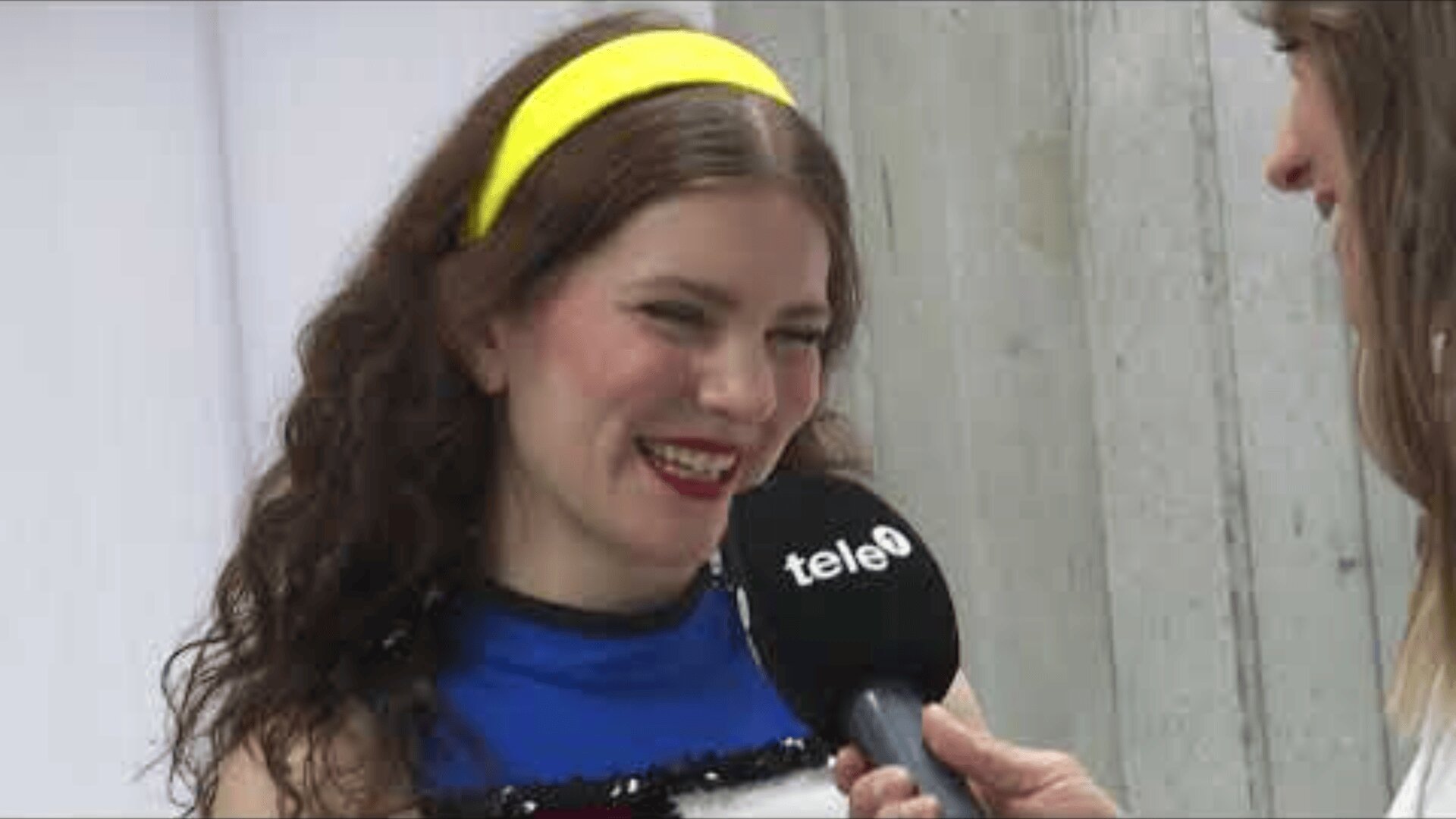 Eine lächelnde Frau mit langen lockigen Haaren und einem gelben Stirnband wird interviewt. Sie spricht in ein schwarzes Mikrofon mit der Aufschrift tele und trägt ein blaues Oberteil. Eine andere Person, die das Mikrofon hält, ist teilweise sichtbar.