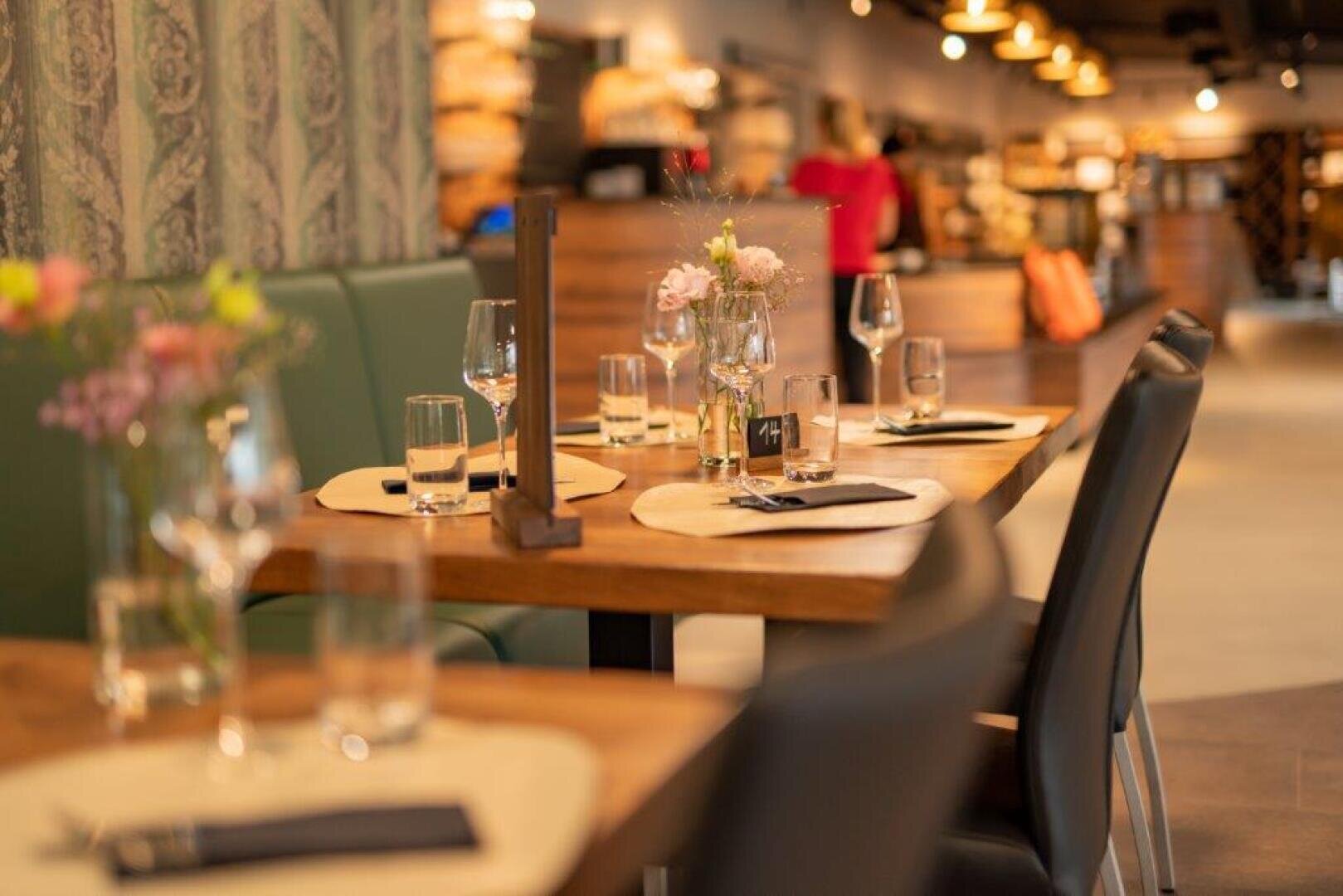 Ein mit Gläsern, Tellern, Besteck und kleinen Blumenvasen gedeckter Restauranttisch; im Hintergrund sind Regale und eine stehende Person zu sehen, die eine warme, einladende Atmosphäre schaffen.