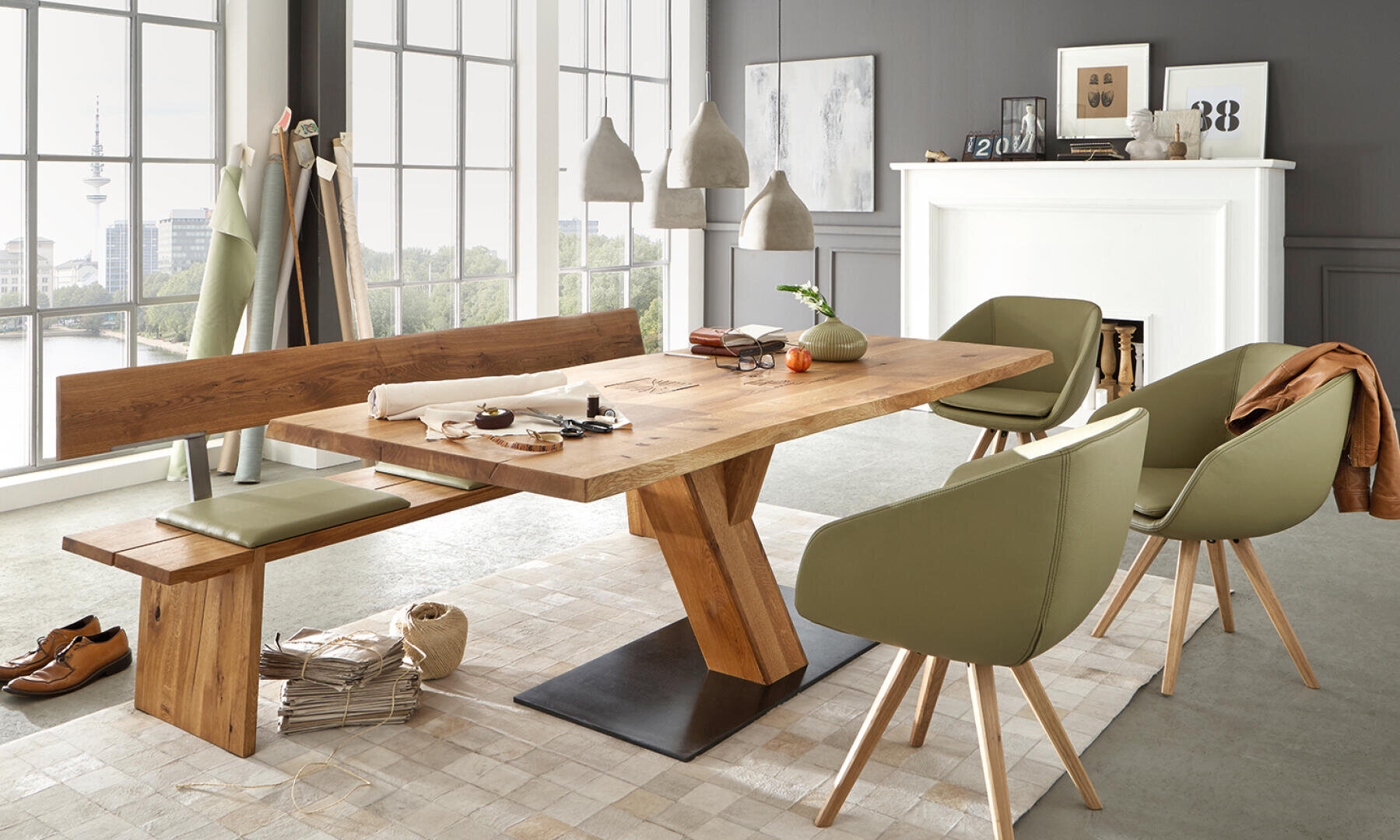 Ein modernes Esszimmer mit einem Holztisch, grün gepolsterten Stühlen und einer passenden Bank. Große Fenster lassen natürliches Licht herein, und die Dekorationsgegenstände sind auf einem weißen Kaminsims und im Raum verteilt.