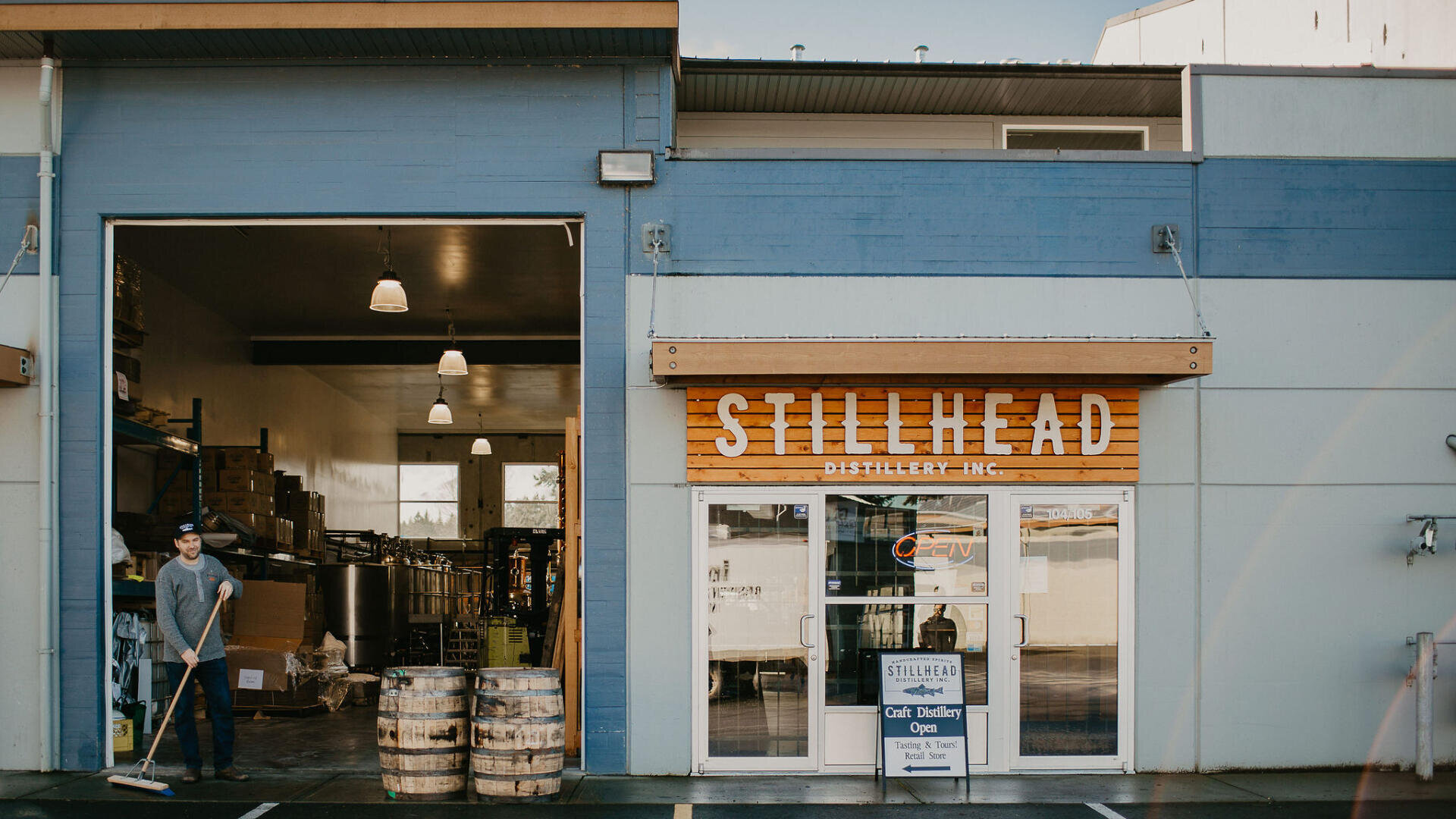 Außenansicht der Stillhead Distillery mit einem Mann, der vor dem offenen Garagentor kehrt; in der Nähe sind Fässer gestapelt, und am Glaseingang sind der Name und die Öffnungszeiten der Brennerei angegeben.