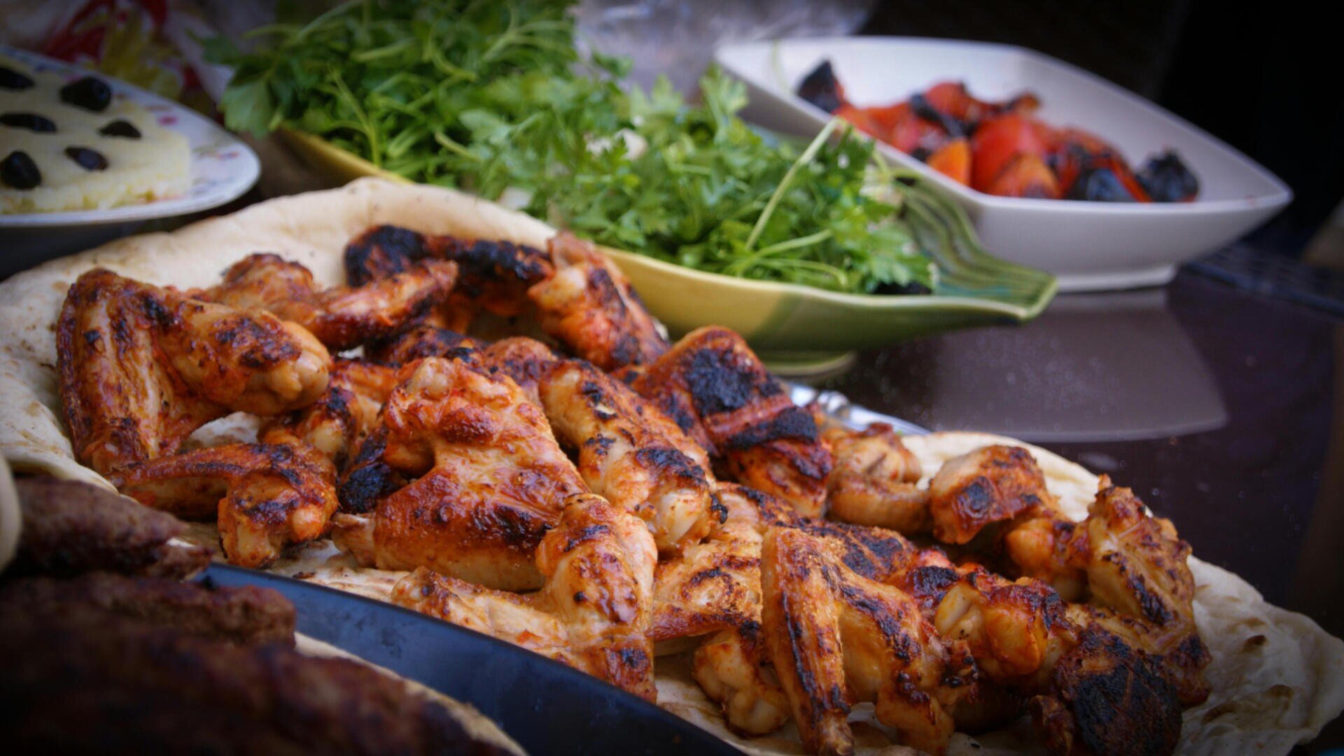 Gegrillte Hähnchenflügel auf Fladenbrot werden mit frischem Grün und einer Schüssel mit gebratenem Gemüse im Hintergrund serviert, wodurch eine farbenfrohe und appetitliche Mahlzeit entsteht.