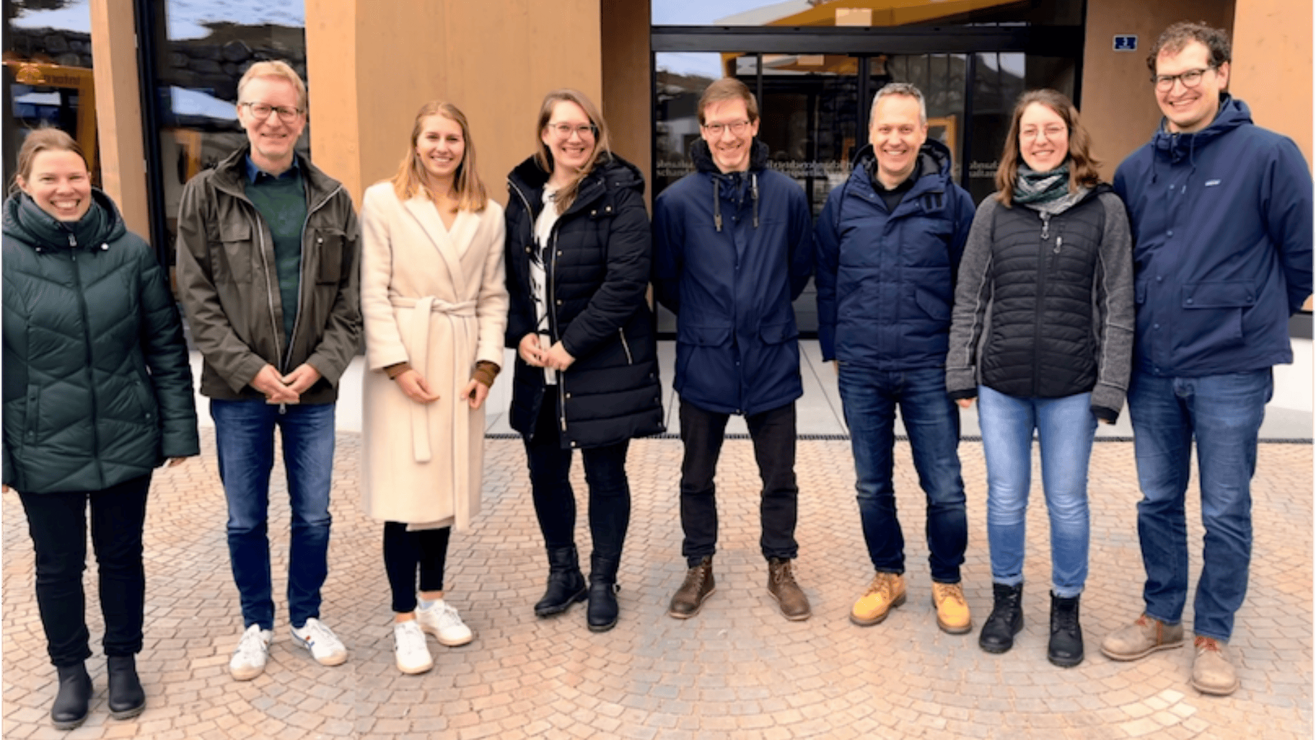 Acht Personen stehen nebeneinander vor einem Gebäude und lächeln in die Kamera. Sie sind in warme Jacken und Winterkleidung gekleidet. Der Boden ist mit gemusterten Steinfliesen bedeckt.