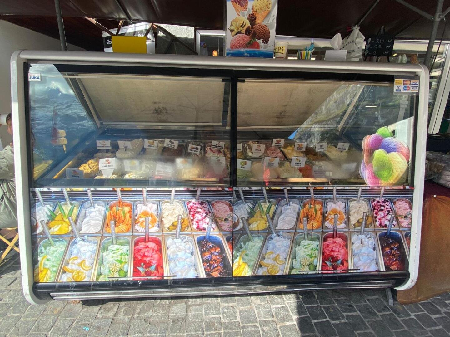 Eine Glasvitrine enthält eine Vielzahl von Käsesorten, die jeweils mit Etiketten versehen sind. Der Boden der Vitrine ist mit einem farbenfrohen Bild von verschiedenen Eissorten und Fruchtbelägen verziert. Im Hintergrund ist die Kulisse des Marktstandes zu sehen.