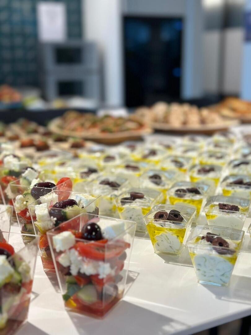 Ein Tisch mit einzelnen durchsichtigen Bechern, die griechischen Salat mit Schafskäse, Oliven und Kirschtomaten enthalten, ordentlich angeordnet für ein Buffet oder ein Catering-Event. Andere Vorspeisen sind im Hintergrund verschwommen.