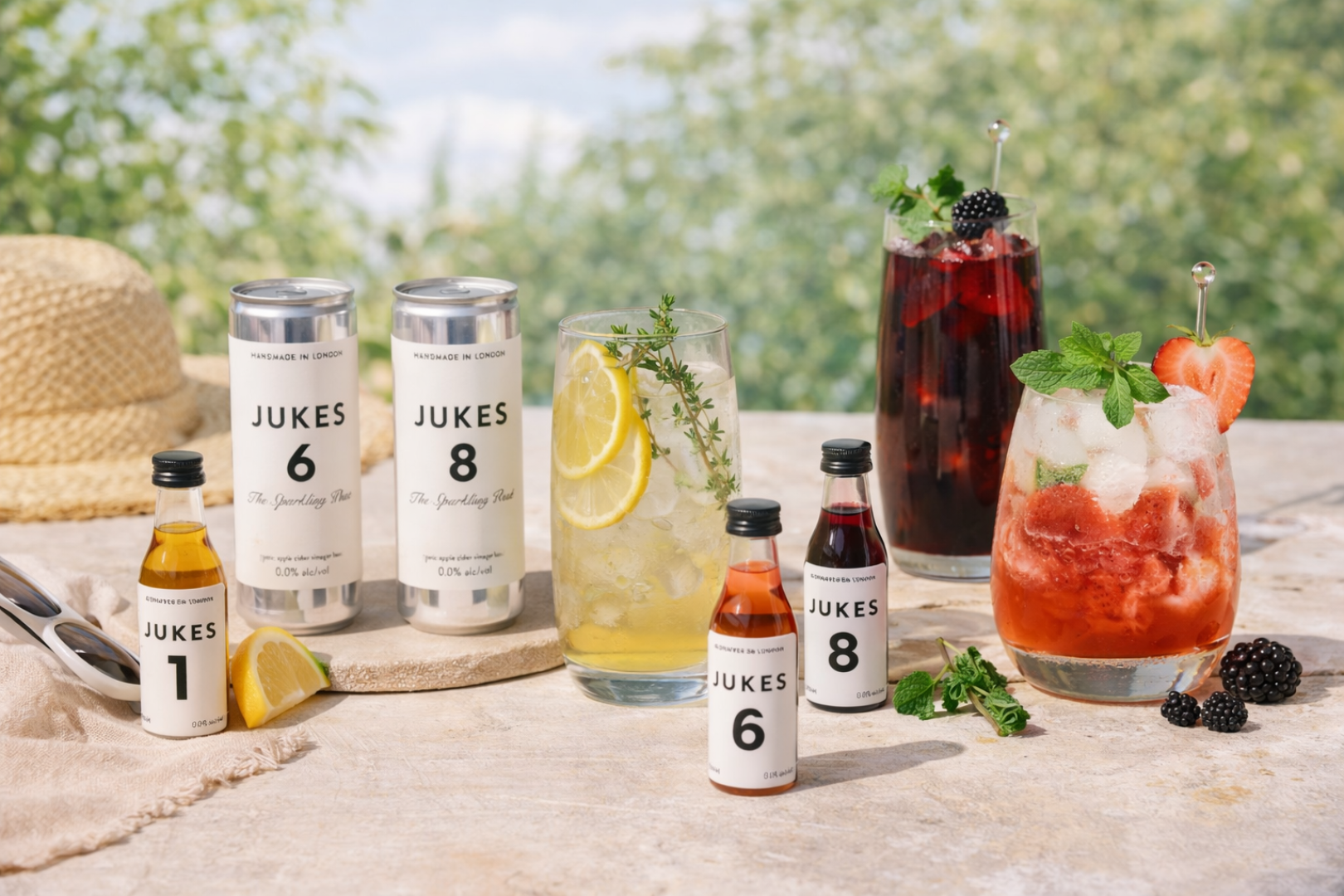 Ein sommerlicher Tisch im Freien mit Jukes-Getränkeflaschen und -dosen, zwei Gläsern mit bunten Cocktails, garniert mit Kräutern und Früchten, einem Sonnenhut, Brombeeren und einem verschwommenen grünen Hintergrund.