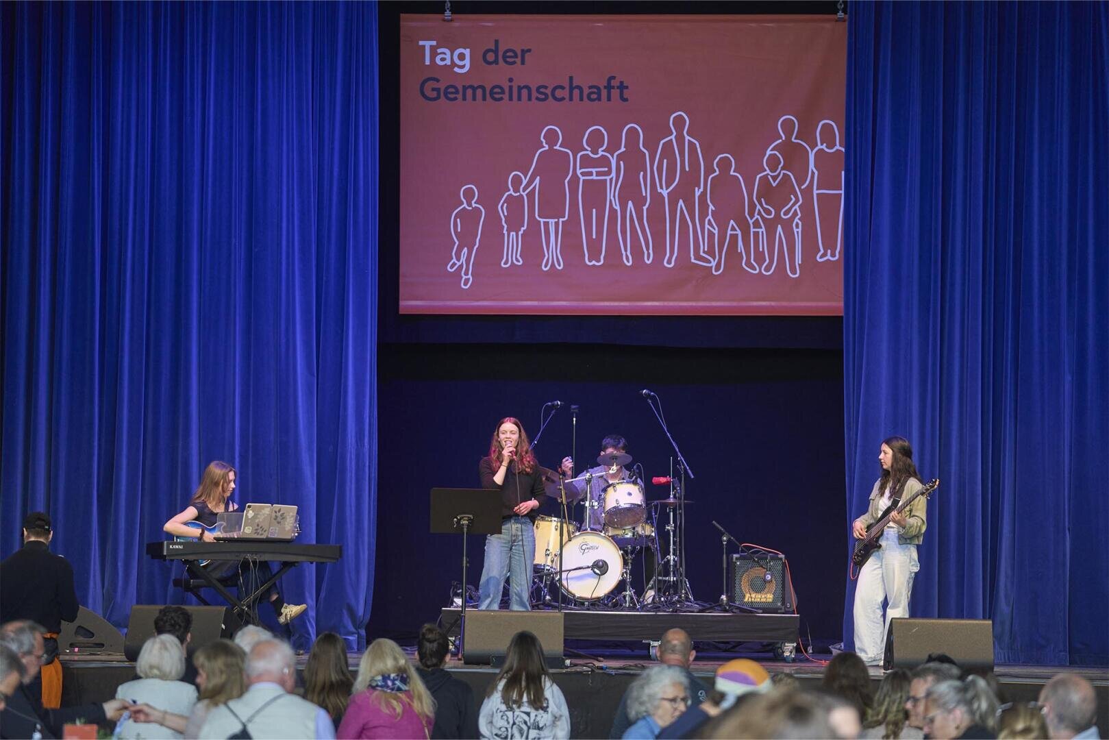 Eine Band mit einem Keyboarder, einem Sänger, einem Schlagzeuger und einem Gitarristen spielt auf der Bühne unter einem roten Banner mit der Aufschrift "Tag der Gemeinschaft", während ein Publikum zusieht. Blaue Vorhänge umrahmen die Bühne.