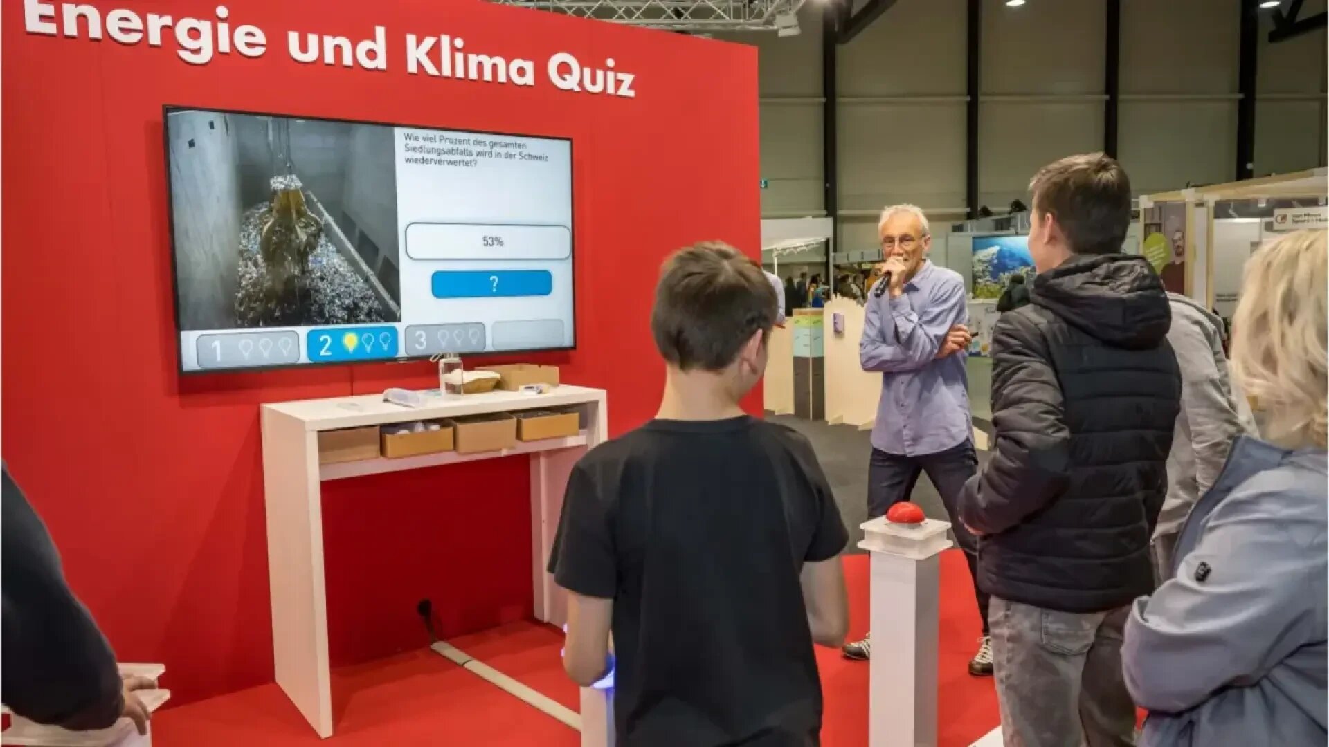 Eine Gruppe von Personen nimmt bei einer Indoor-Veranstaltung an einem Energie- und Klima-Quiz teil. Sie stehen vor einem großen Bildschirm, auf dem eine Multiple-Choice-Frage und die prozentualen Ergebnisse angezeigt werden; ein Mann mit einem Mikrofon moderiert.