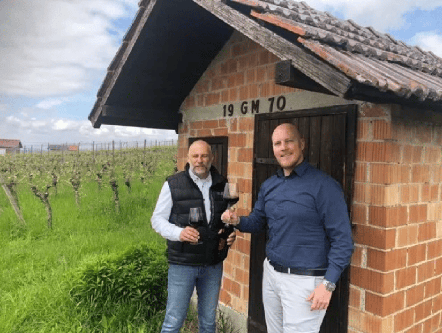 Zwei Männer stehen vor einem kleinen Backsteingebäude mit der Aufschrift 19 GM 70, halten Gläser mit Rotwein in der Hand und lächeln, während im Hintergrund Weinreben und ein bewölkter Himmel zu sehen sind.