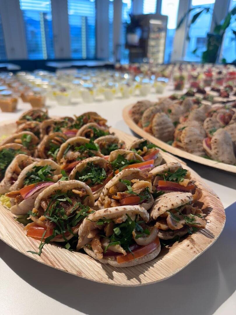 Eine Nahaufnahme von zwei Platten mit Mini-Pita-Sandwiches, gefüllt mit Gemüse und Kräutern, auf einem Tisch, mit verschiedenen Vorspeisen und Desserts im Hintergrund.