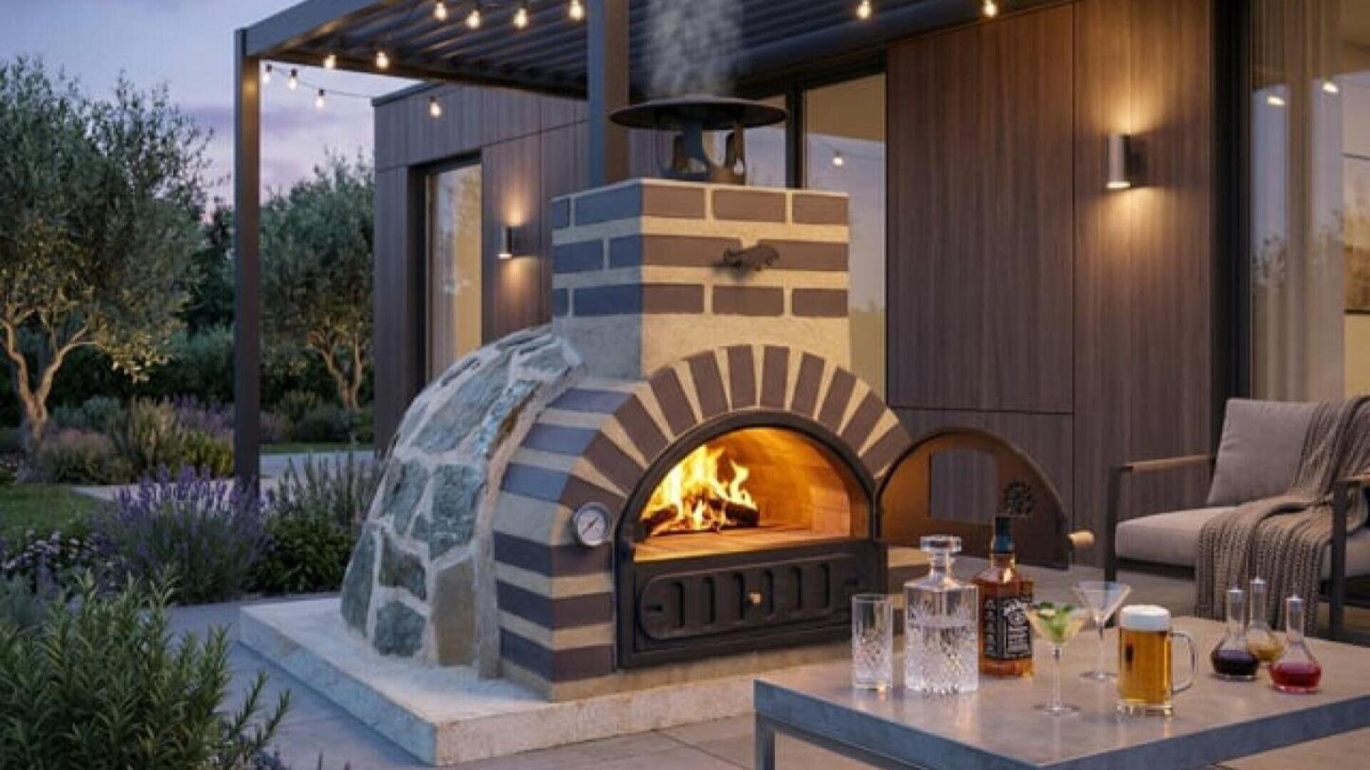 Außenterrasse mit modernem holzbefeuertem Ziegelofen, Feuer im Inneren, überdachter Pergola, gemütlichen Sitzgelegenheiten, Lichterketten und einem Tisch mit Getränken und Gläsern im Vordergrund.