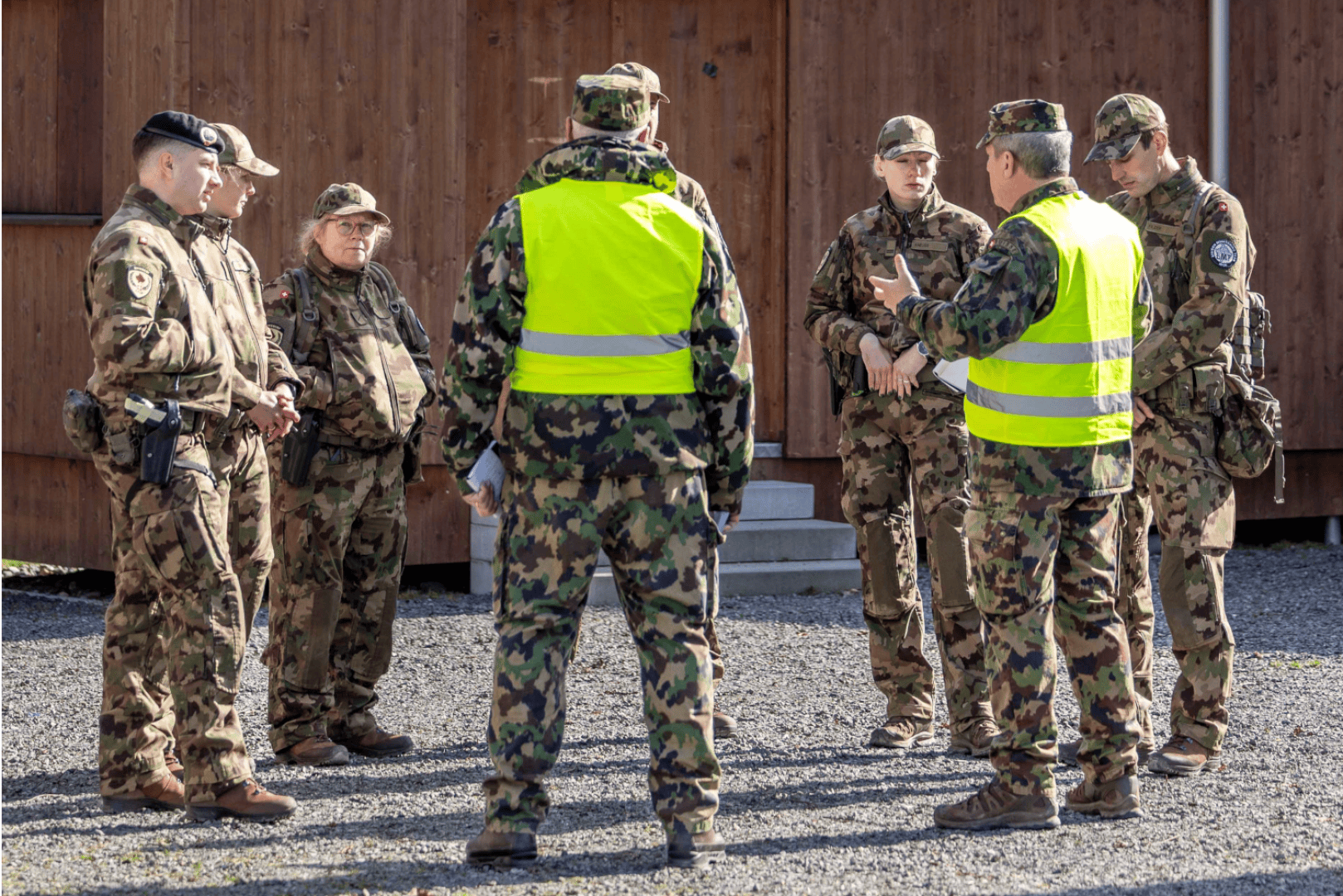 Eine Gruppe von Soldaten in Tarnuniformen und gelben Sicherheitswesten steht in einem Kreis im Freien und diskutiert vor einem Holzgebäude.