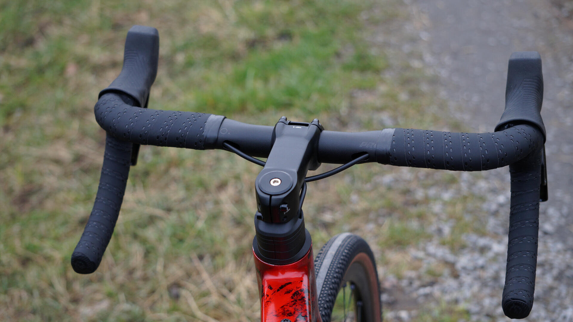 Nahaufnahme eines schwarzen Lenkers an einem roten Fahrrad, das auf einer Wiese neben einem Kiesweg steht.