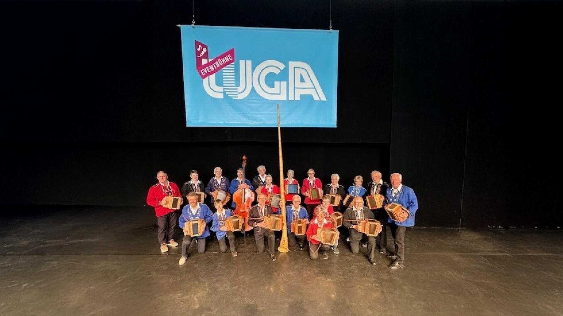 Eine Gruppe von Musikern steht auf der Bühne und hält Konzertinas in der Hand. Hinter ihnen hängt ein großes blaues Banner mit der Aufschrift CUGA. Sie tragen bunte Hemden, und eine Person hält ein großes Alphorn in der Mitte.